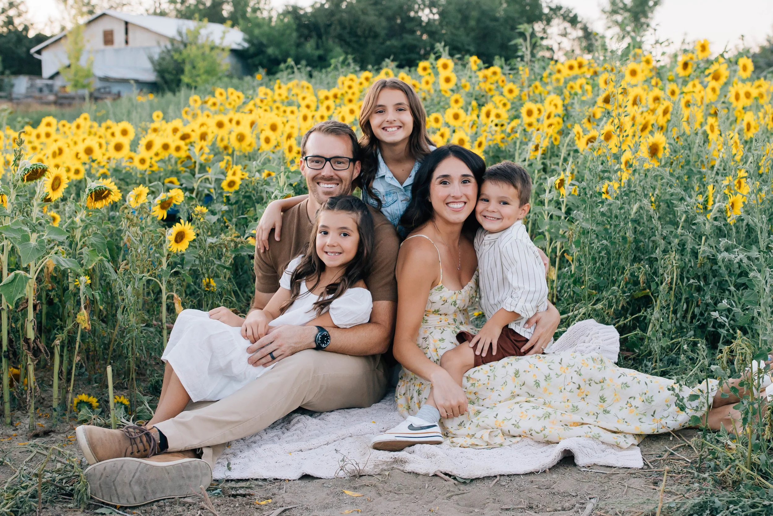 A family of five sitting on a blanket in a sunflower field, smiling at the camera. The group includes two adults and three children, with a barn visible in the background.