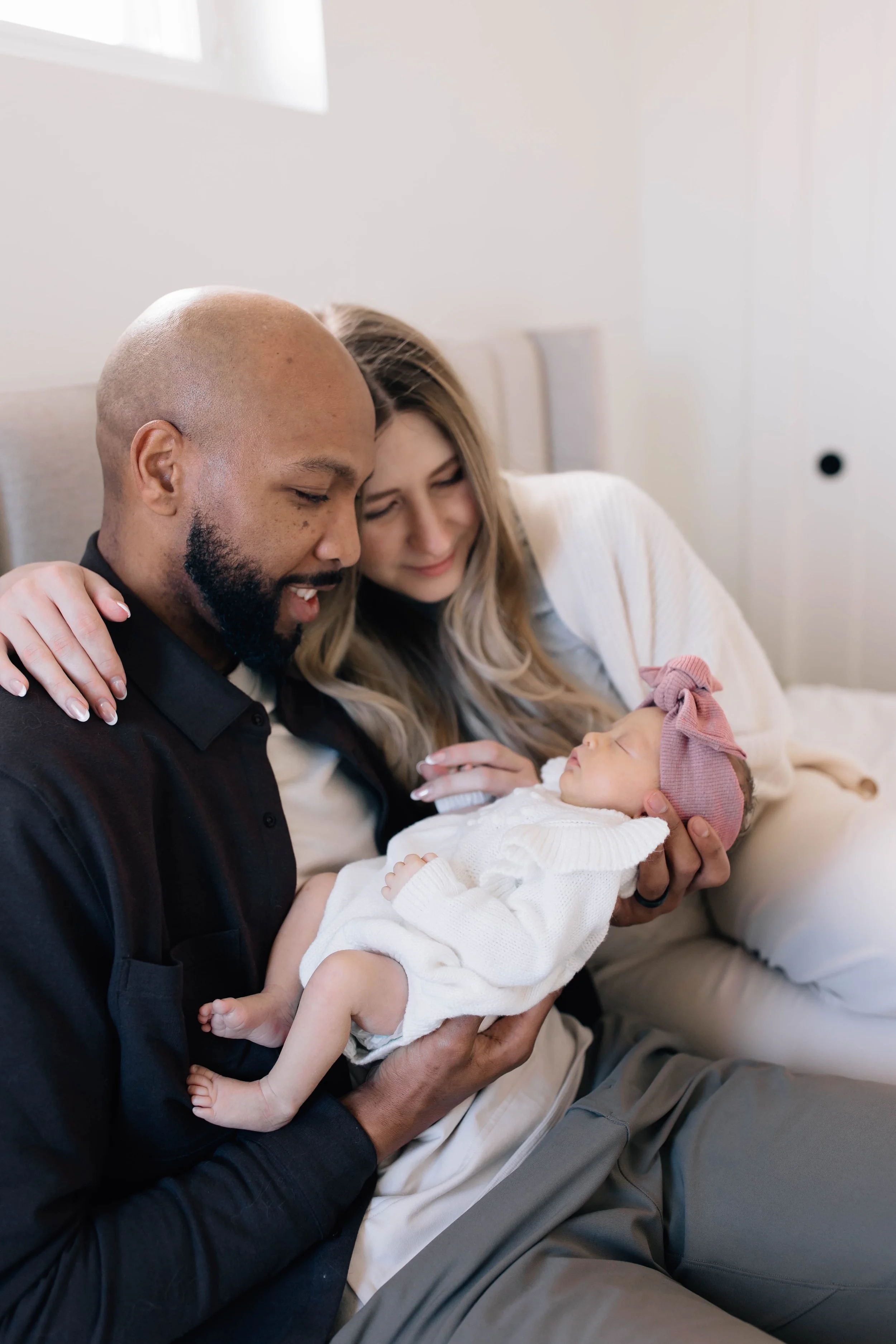 A man and woman sitting on a bed, holding a sleeping baby girl dressed in white with a pink bow headband, smiling and looking at the baby.