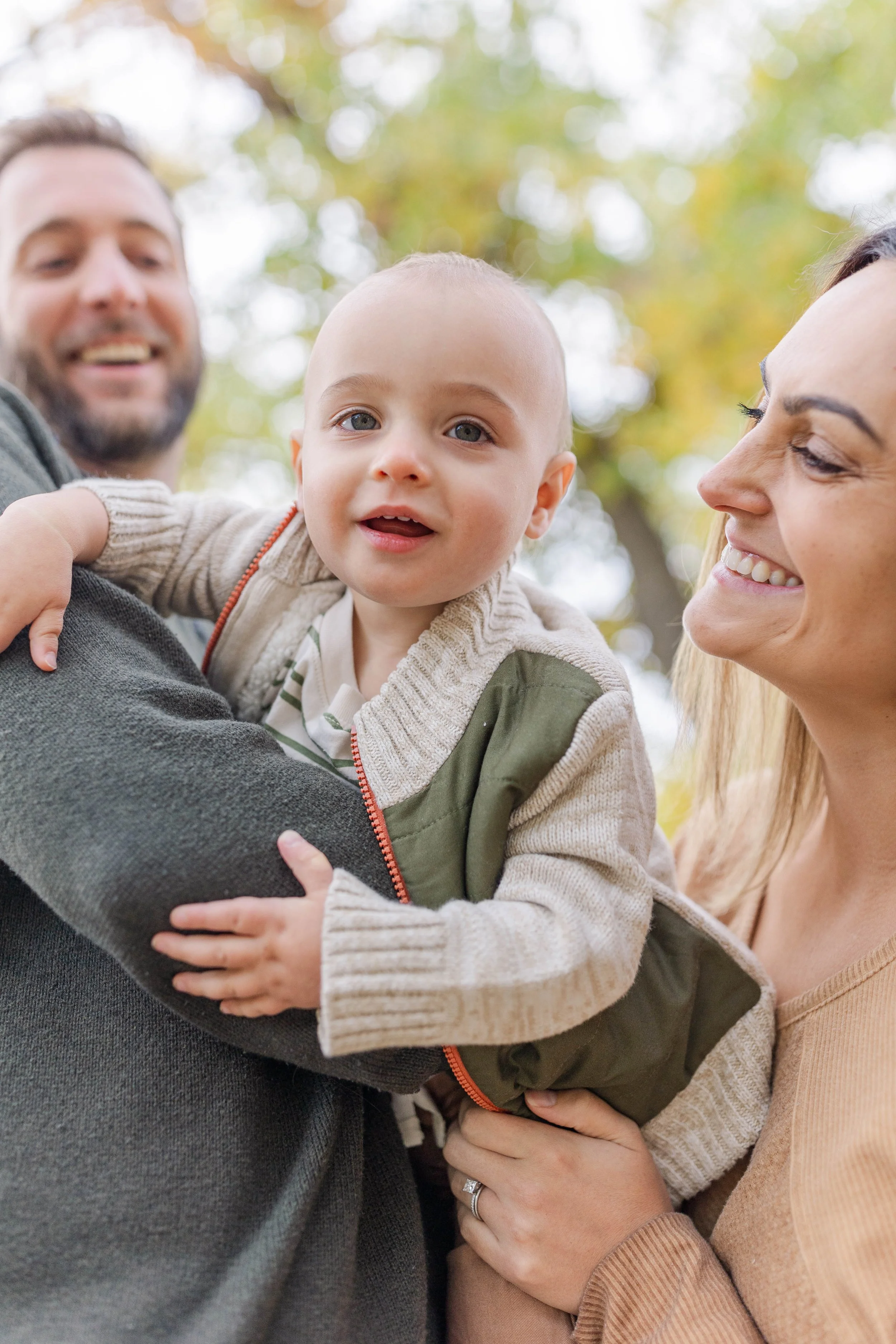 A happy family outdoors, with a smiling woman and man holding their young child between them, surrounded by trees with autumn leaves.