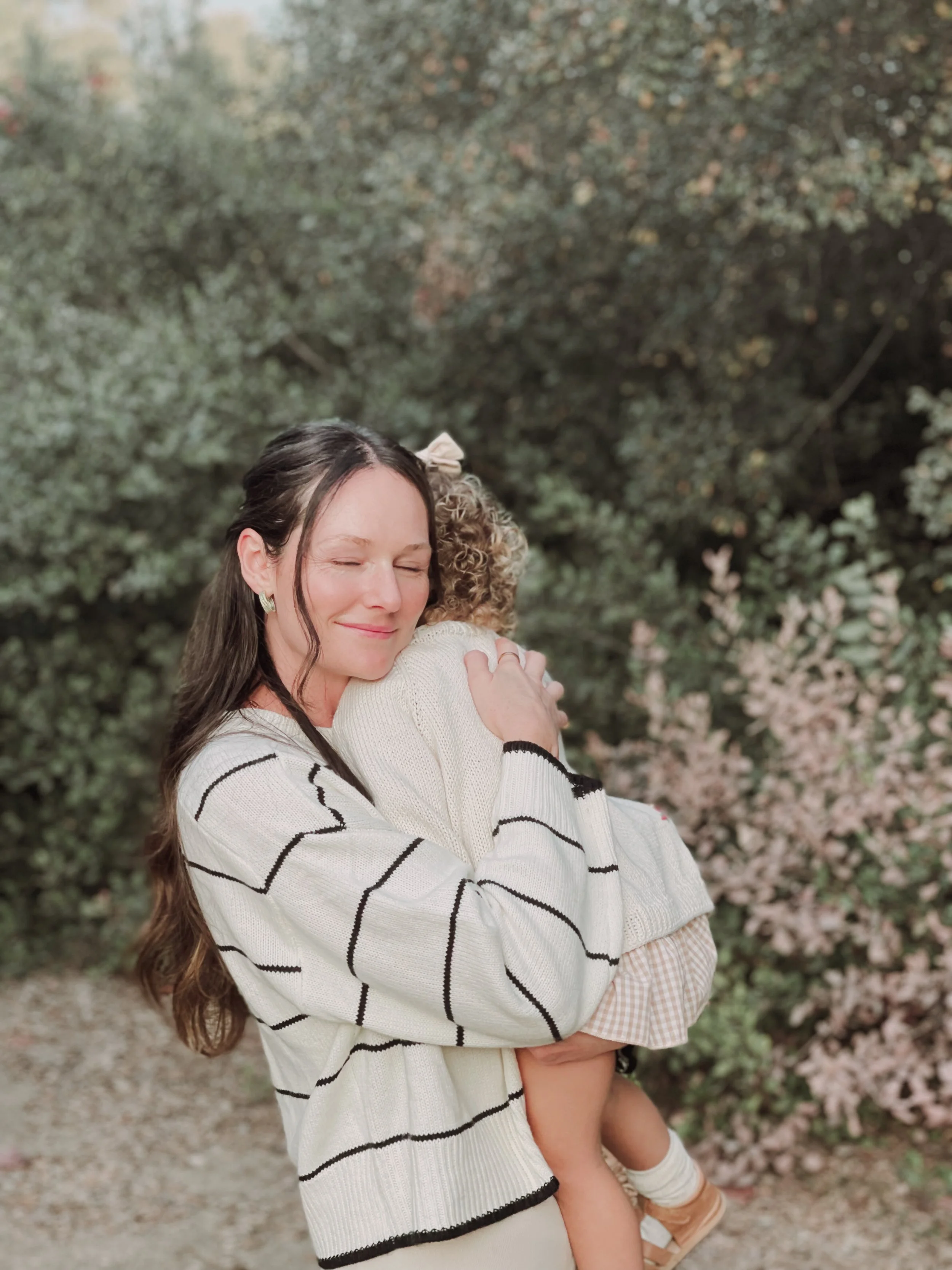A woman with long brown hair and a white sweater with black stripes holding a young girl with curly hair in a beige sweater outdoors surrounded by greenery and blooming flowers.