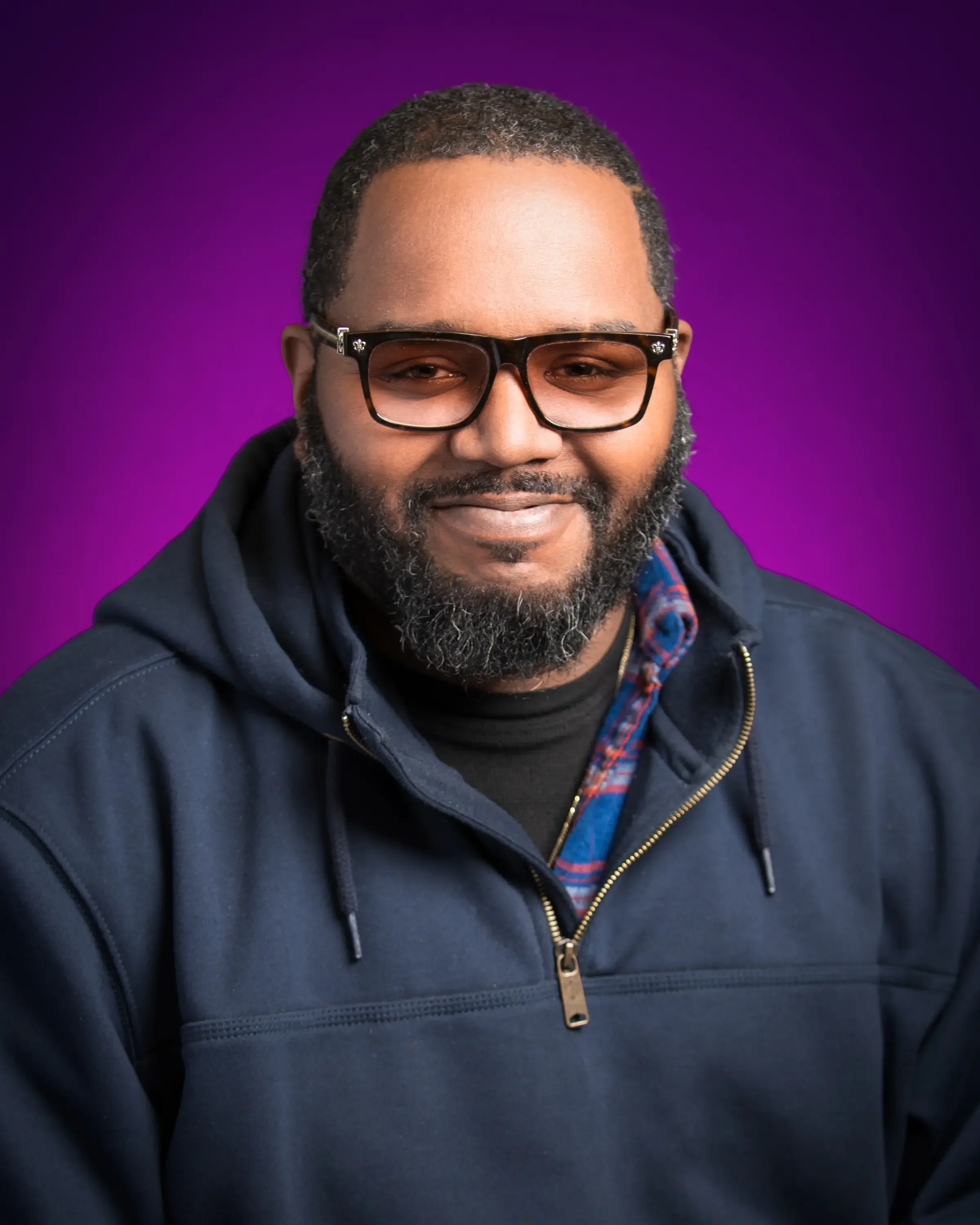Portrait of a smiling man with glasses, a beard, wearing a dark hoodie and a plaid shirt, against a purple background.