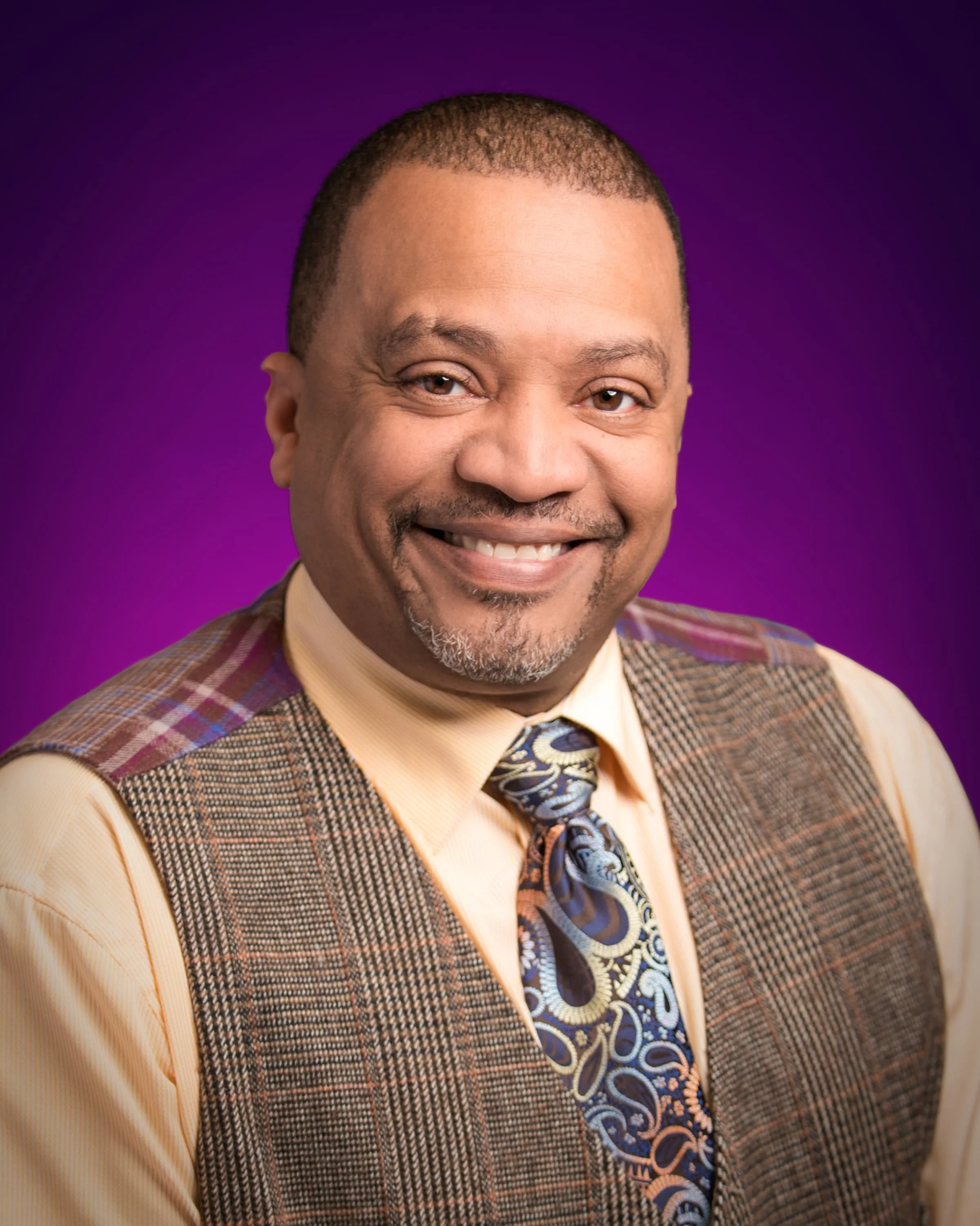 A smiling man with short hair, wearing a yellow shirt, patterned vest, and a colorful patterned tie, against a purple background.