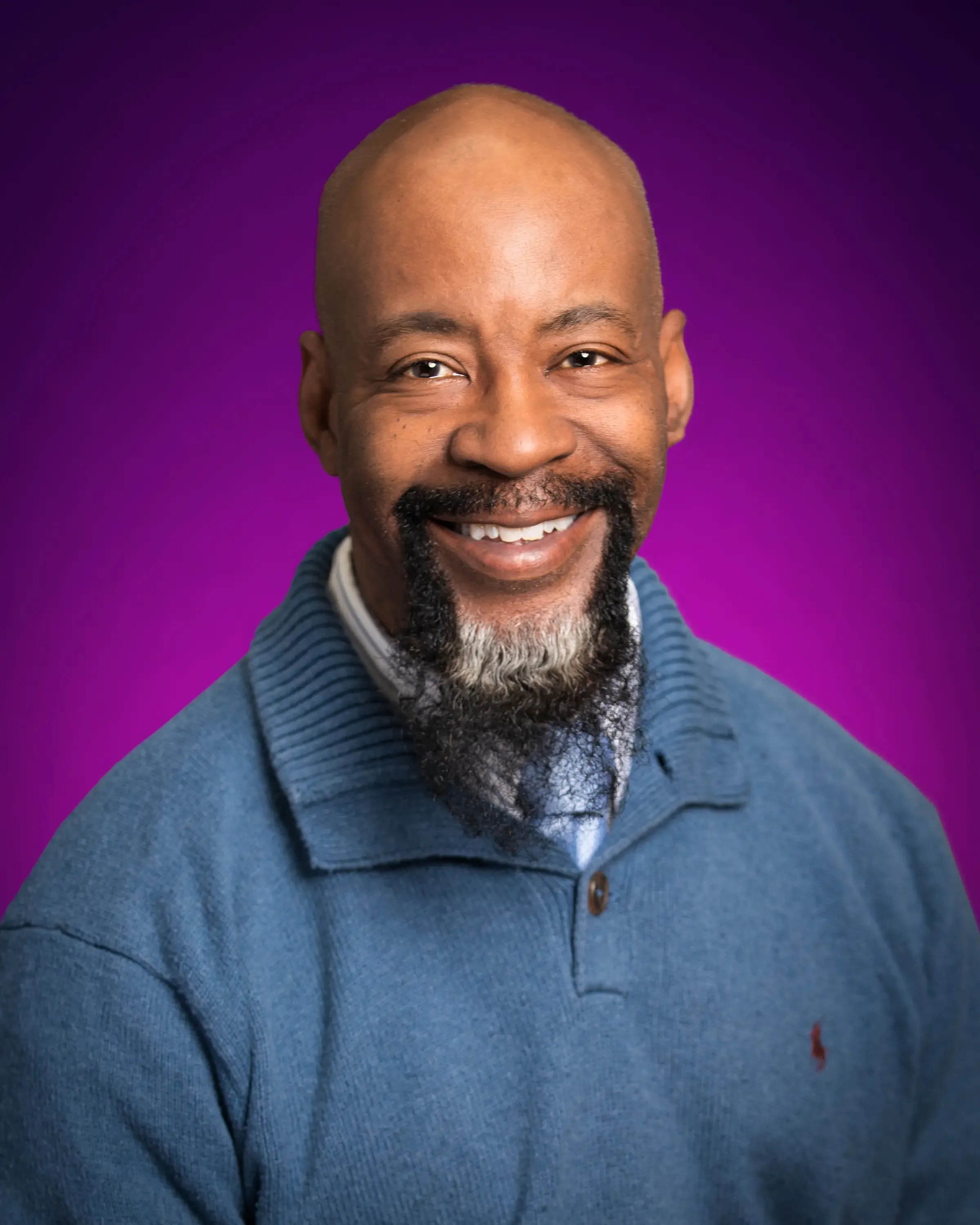 Headshot of a smiling african-american man with a bald head, beard and mustache, wearing a blue sweater, against a purple background.