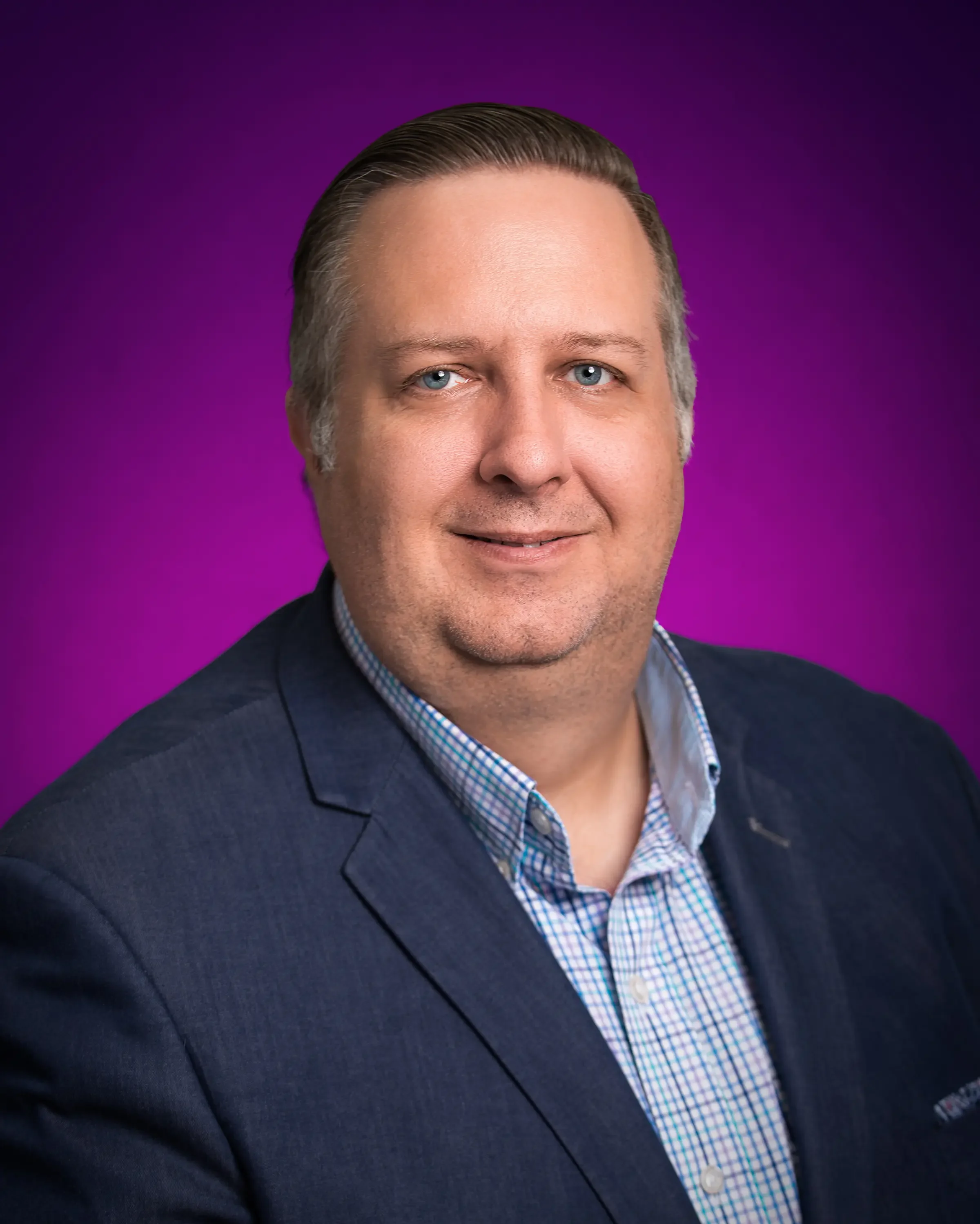 Headshot of a man with light skin, short dark blond hair, wearing a dark blazer and a checkered shirt, smiling against a purple background.