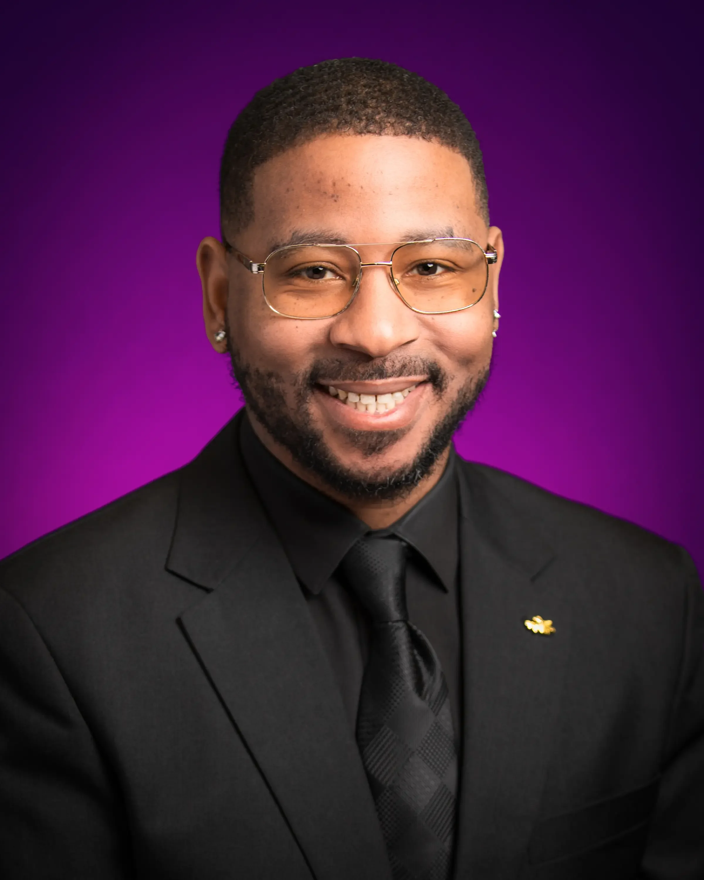 A man wearing glasses, a black suit, and a black tie smiling against a purple background.