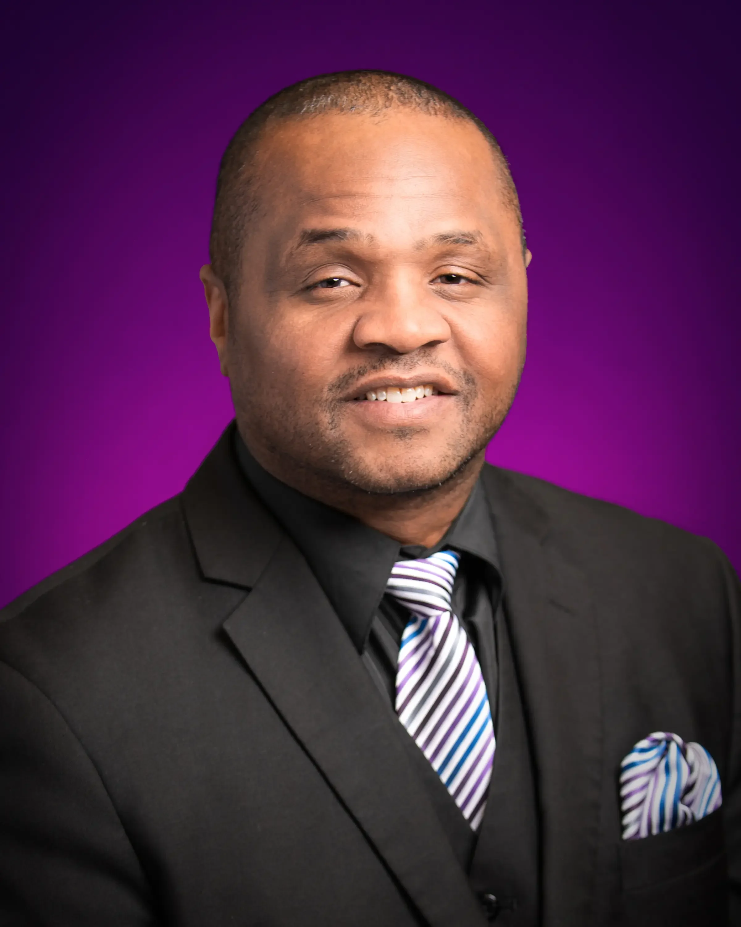 A professional man in a black suit, striped tie, and matching pocket square posing against a purple gradient background.