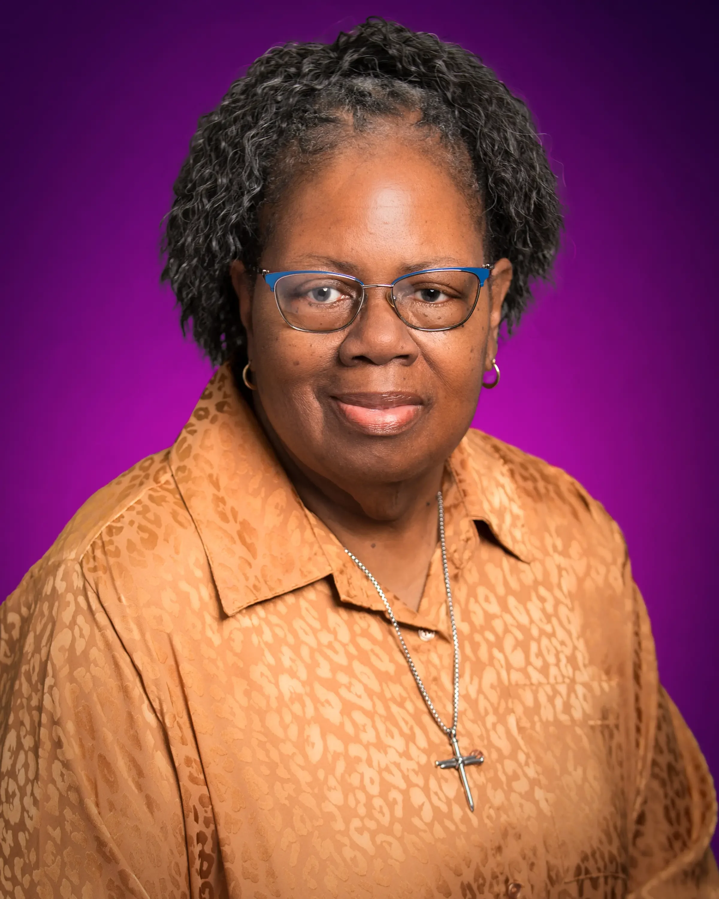 Portrait of a woman with short, curly black hair, wearing glasses, a leopard print shirt, and a silver cross necklace, against a purple background.