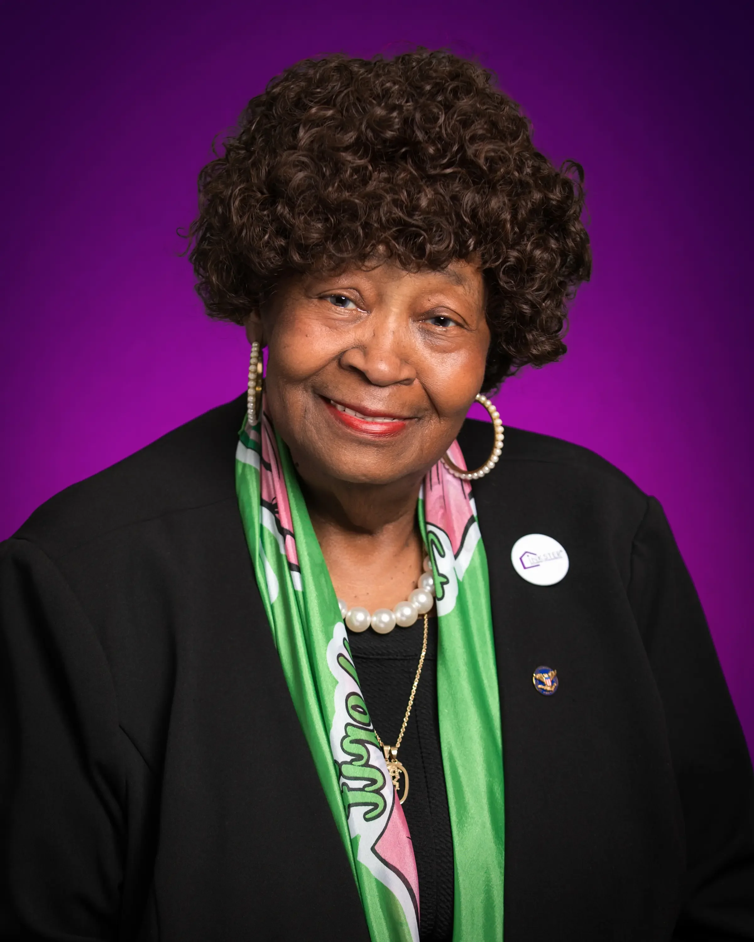 Portrait of an elderly woman with curly hair, wearing a black jacket with pins, a pearl necklace, gold chain, and a green scarf, against a purple background.