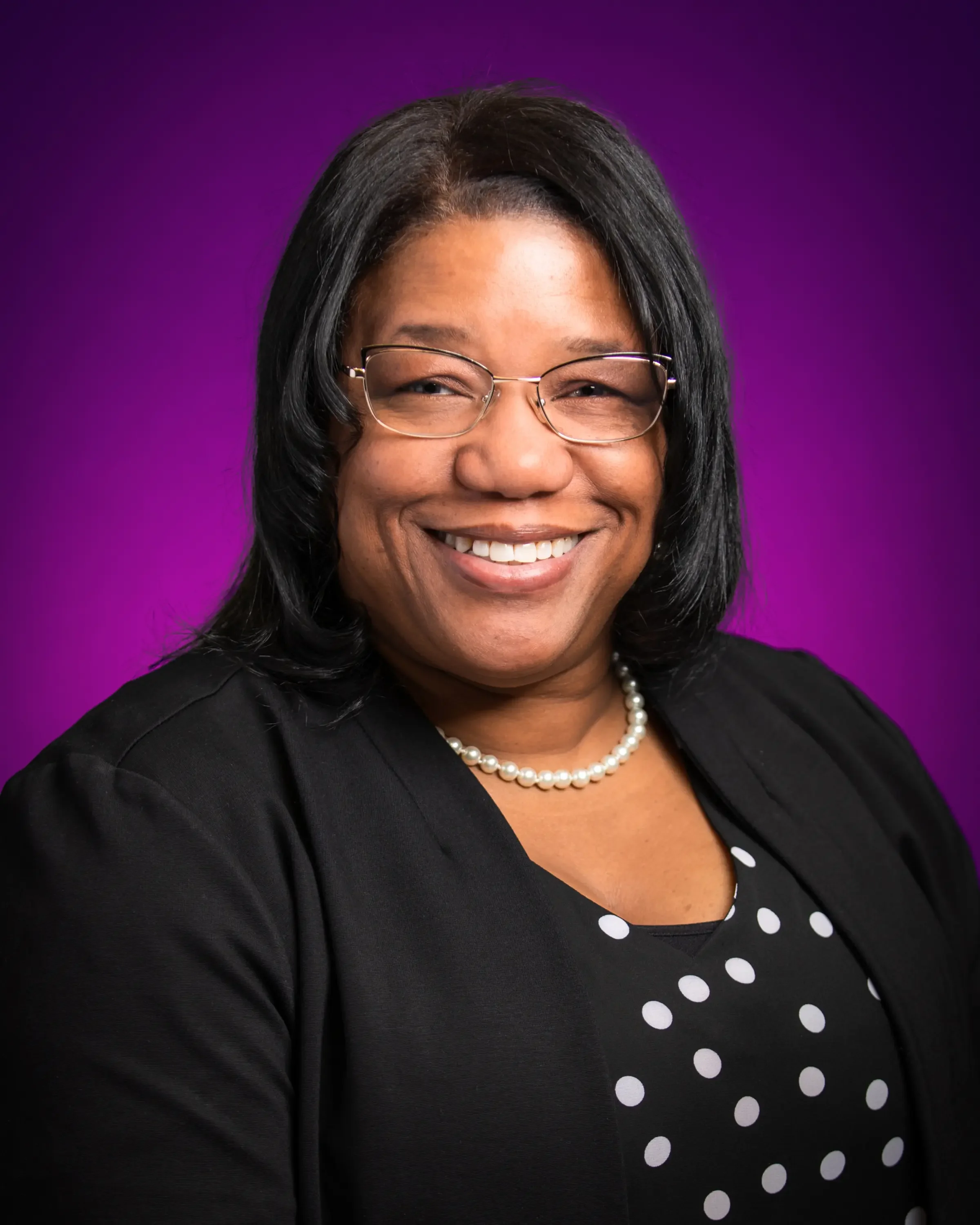 A professional woman with glasses, wearing a black blazer and a polka dot blouse, smiling against a purple background.