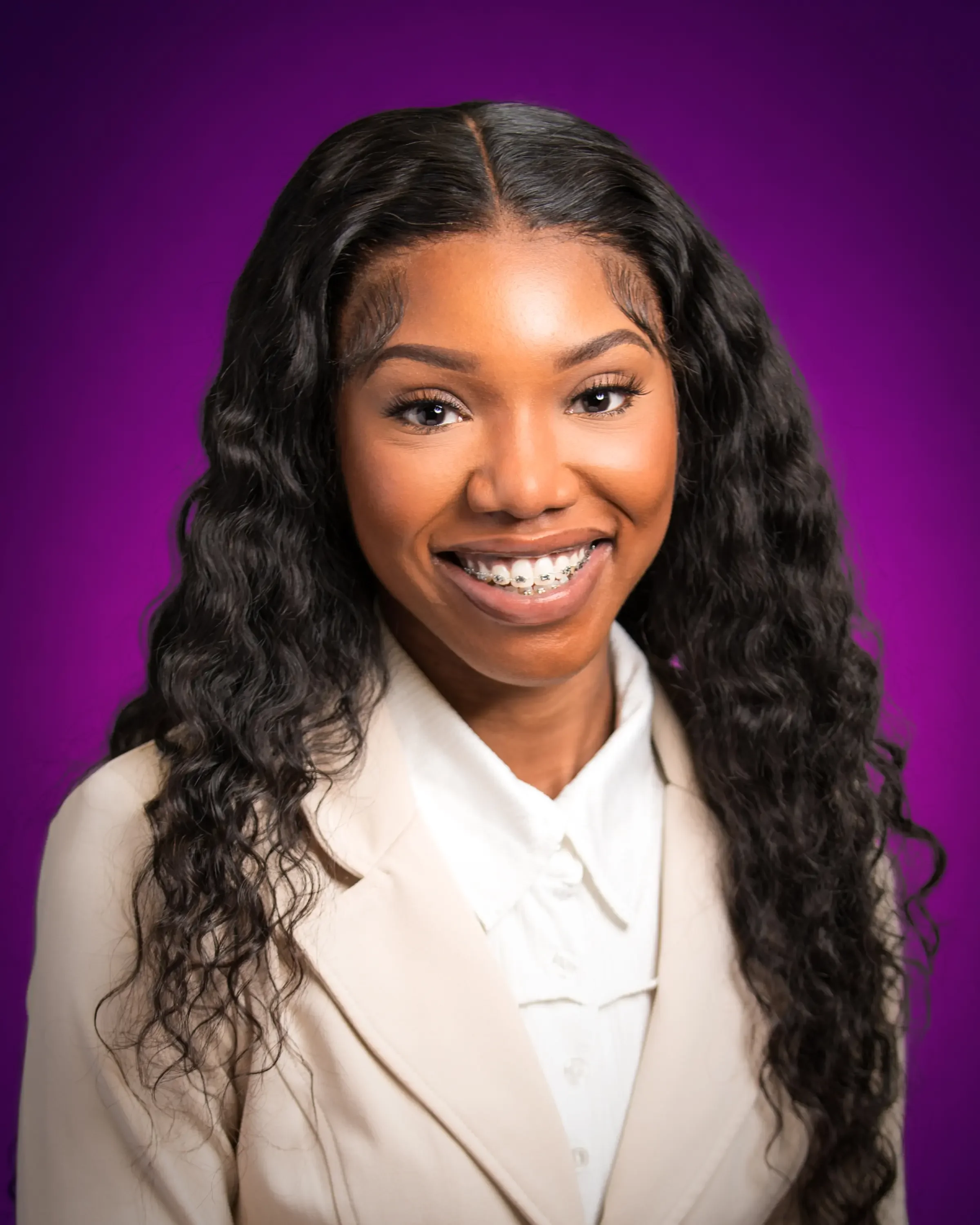 Portrait of a smiling young woman with long curly black hair, wearing a cream blazer and white shirt, against a purple background.
