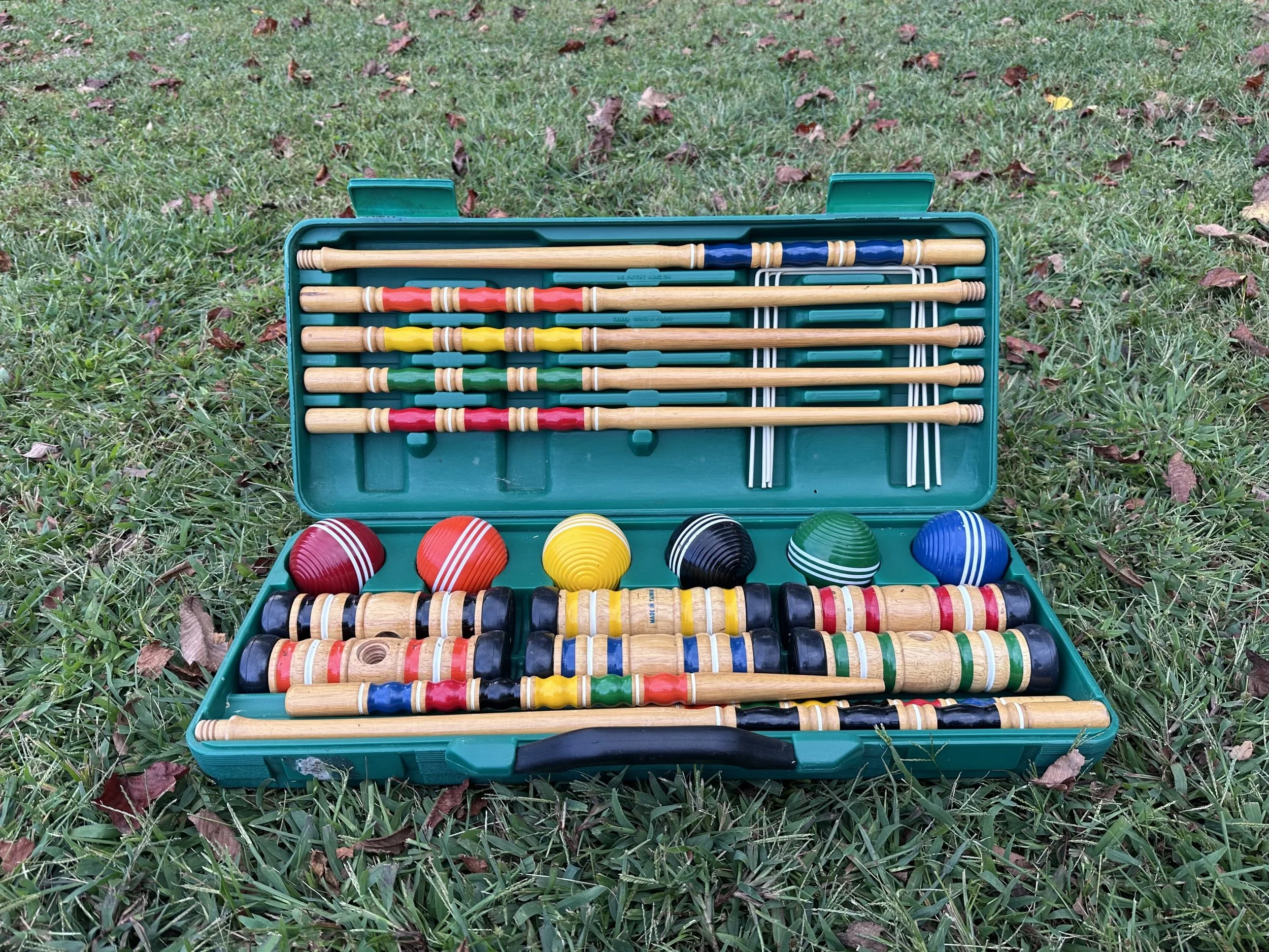 A set of lawn game equipment, including wooden croquet mallets, colored balls, and metal stakes, inside a blue plastic case on grass.