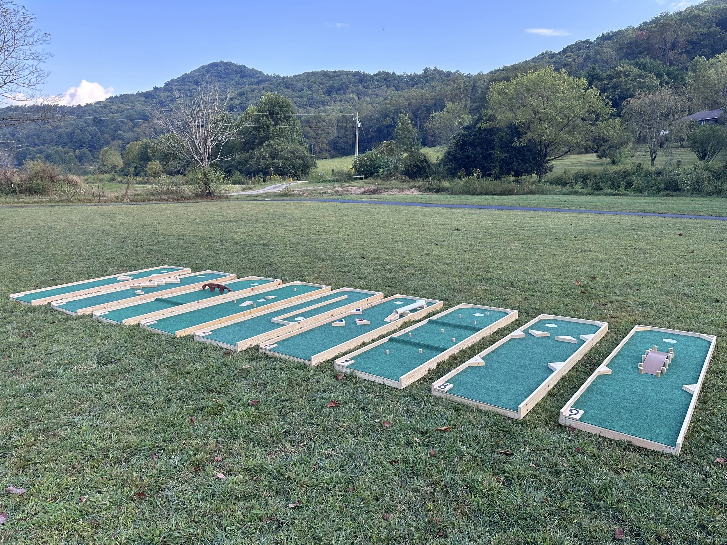 Multiple mini golf holes set up on a grassy field in a rural area, with trees, hills, and a blue sky in the background.