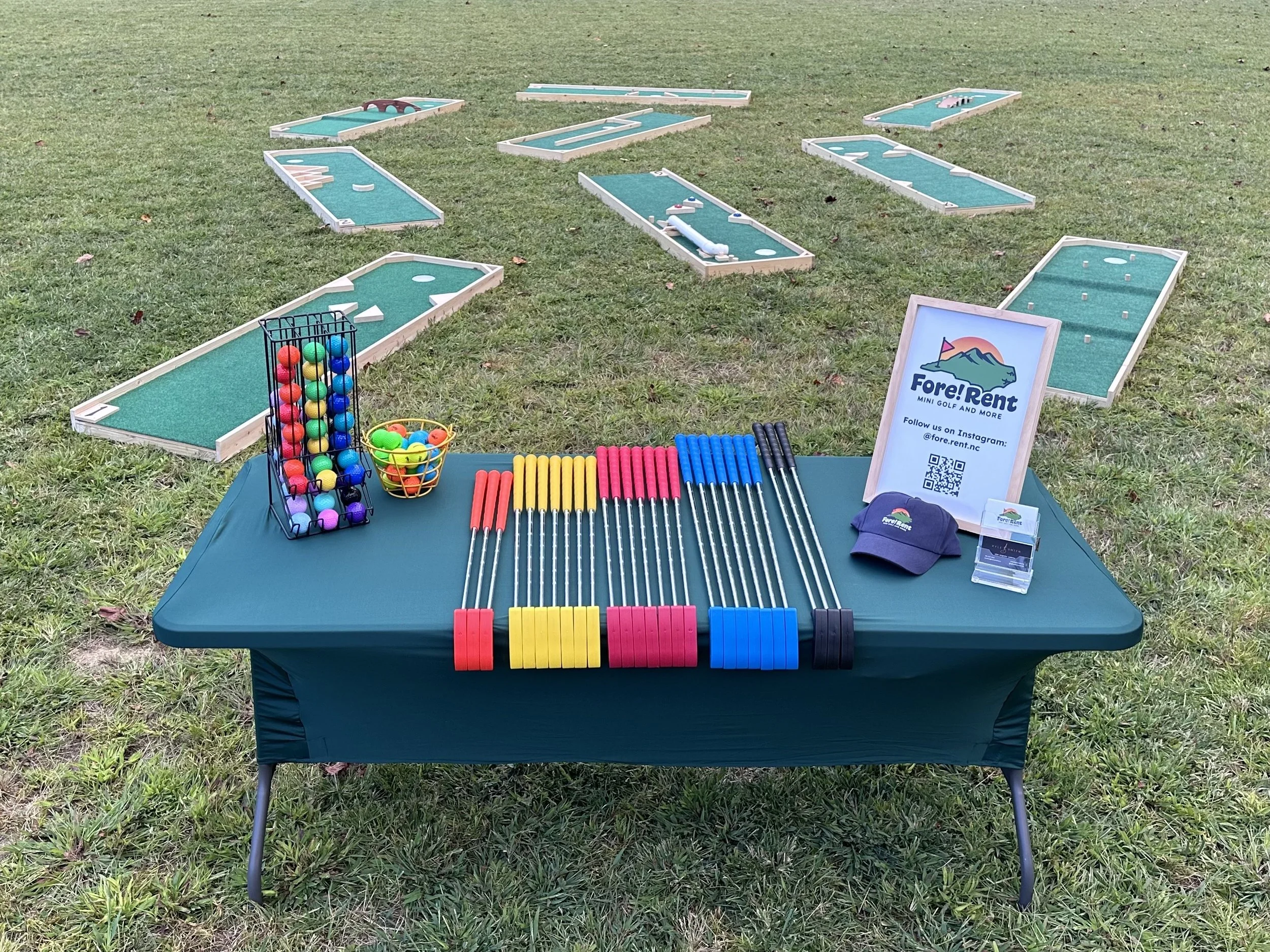 Mini golf setup with colorful putters, balls, and obstacles, outdoors on grass, with a table displaying promotional materials and a sign advertising Fore Rent mini golf.