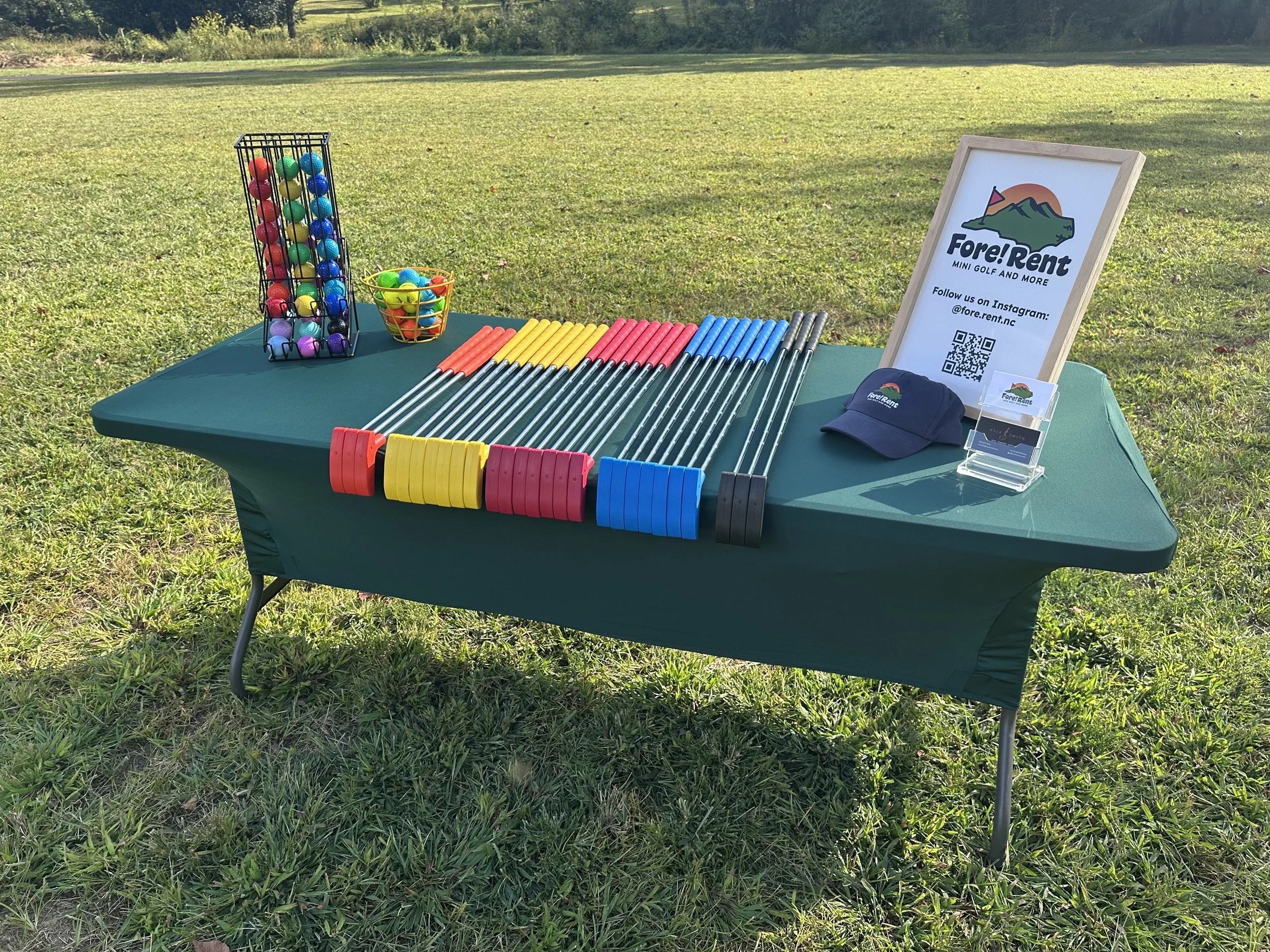 A table set up outdoors with mini golf equipment, including colorful golf balls, putters, and a sign for Fore! Rent mini golf and more, with a QR code and Instagram handle.