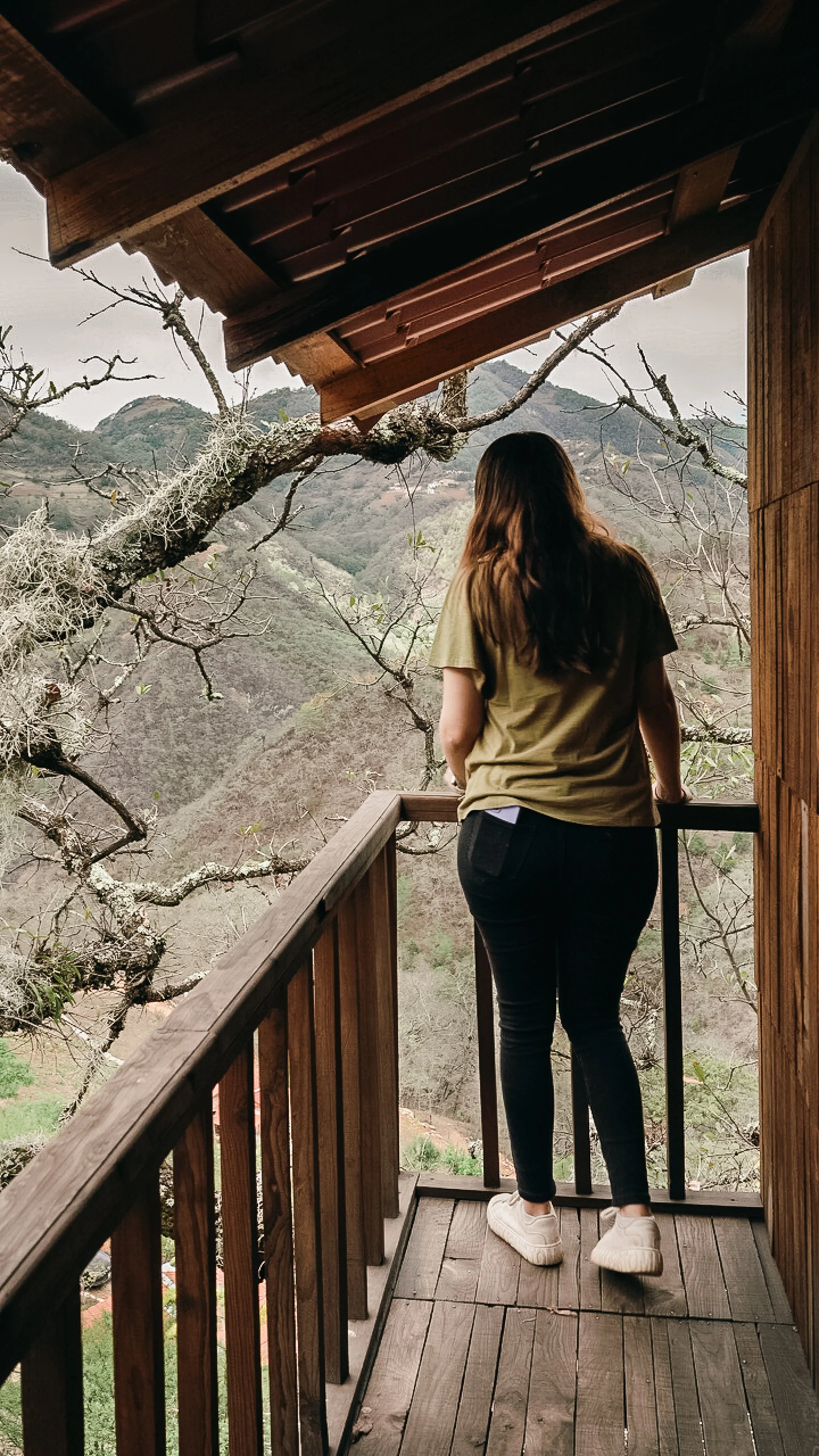 Mujer de espaldas en una terraza de madera, mirando un paisaje montañoso con árboles desnudos y en matorrales.