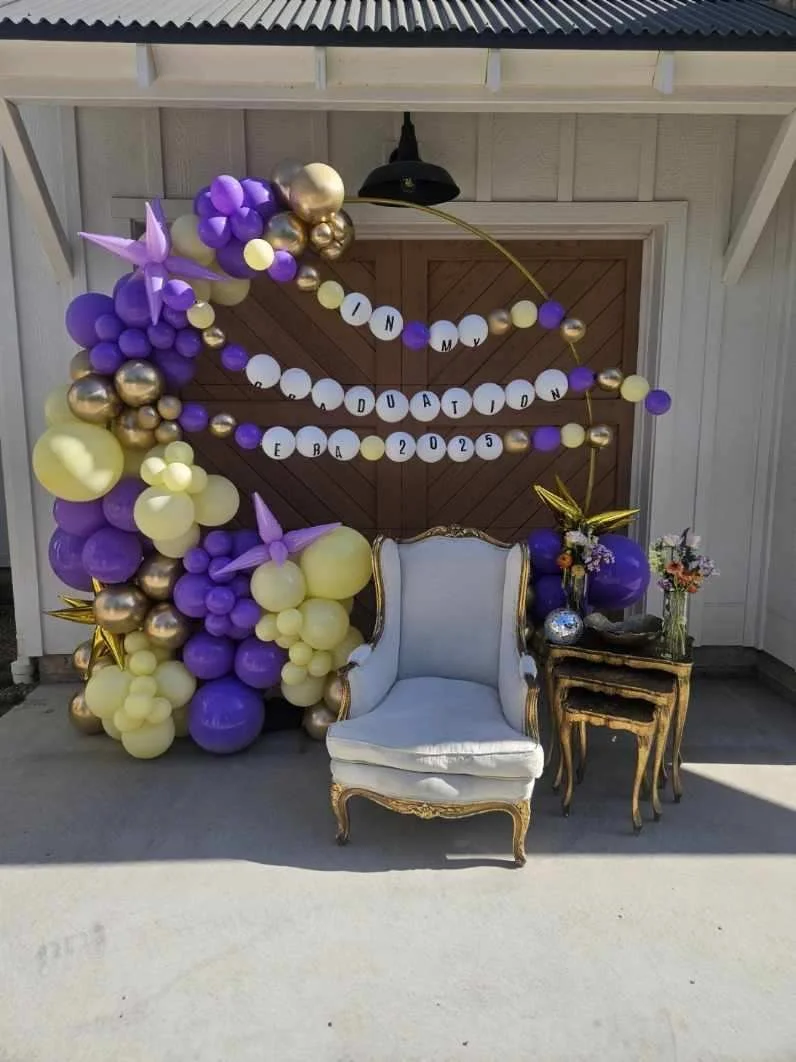 Decorative balloon arch and garland with purple, gold, and white balloons, including gold star balloons, behind a white vintage armchair and a small black and gold side table with flowers, celebrating a graduation event in 2025.
