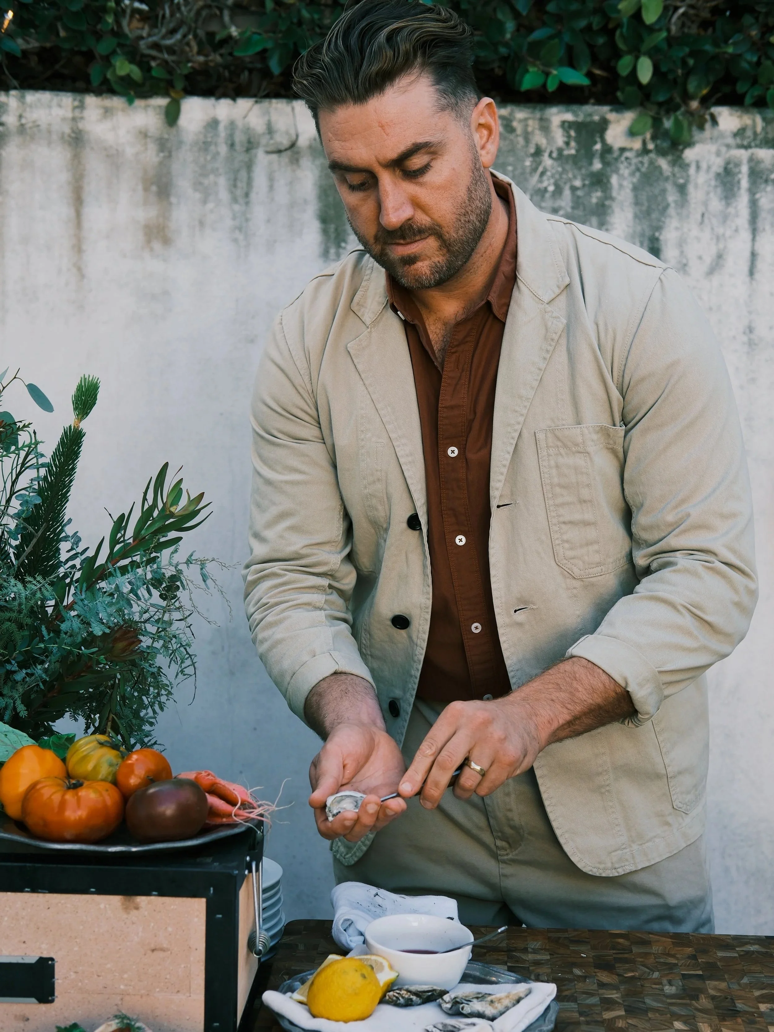 A man preparing seafood outdoors next to a table with fresh vegetables, lemon, and seafood, with greenery and a white wall in the background.