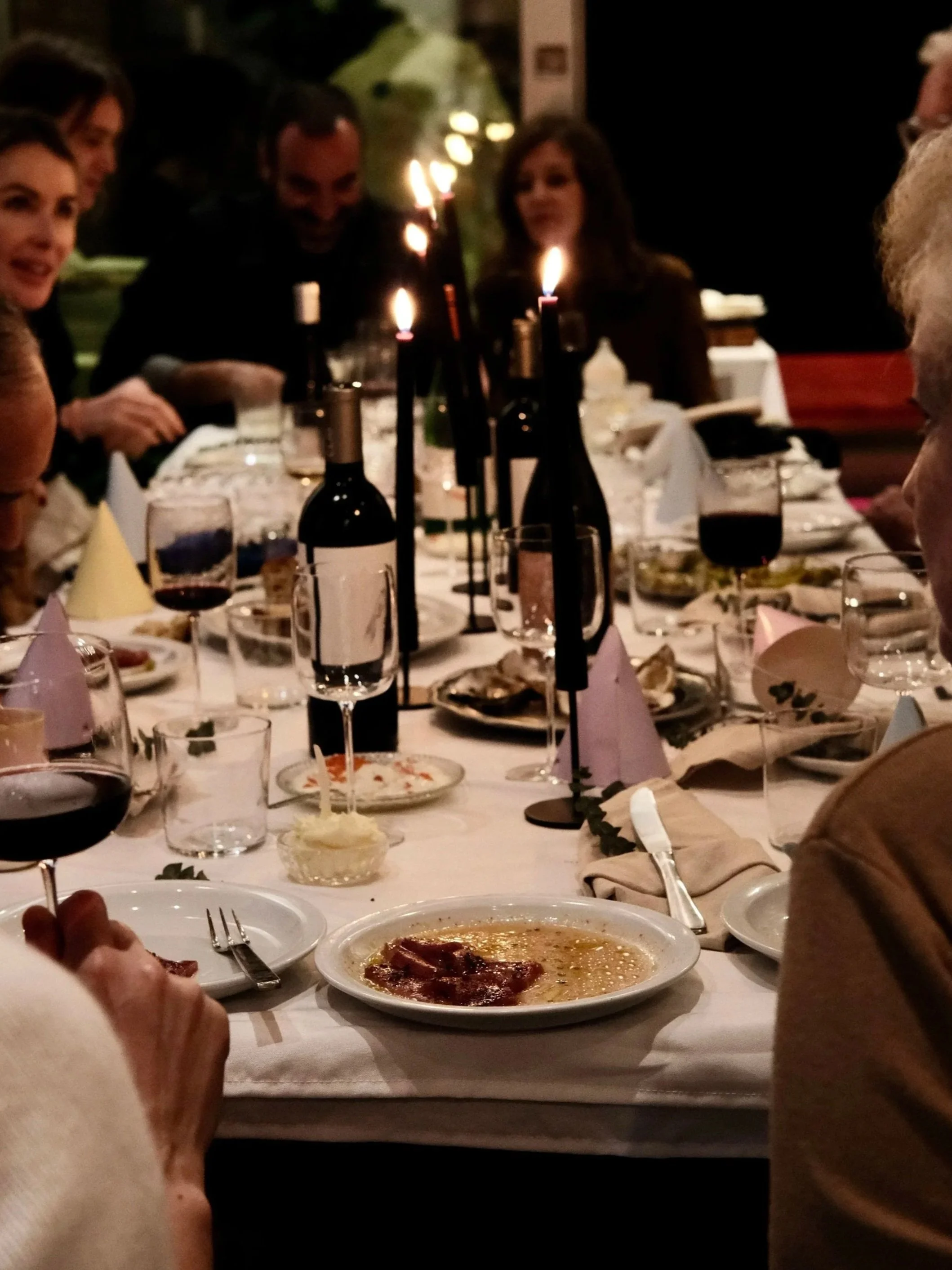 People gathered around a dinner table with candles and wine bottles, enjoying a meal at a social gathering.