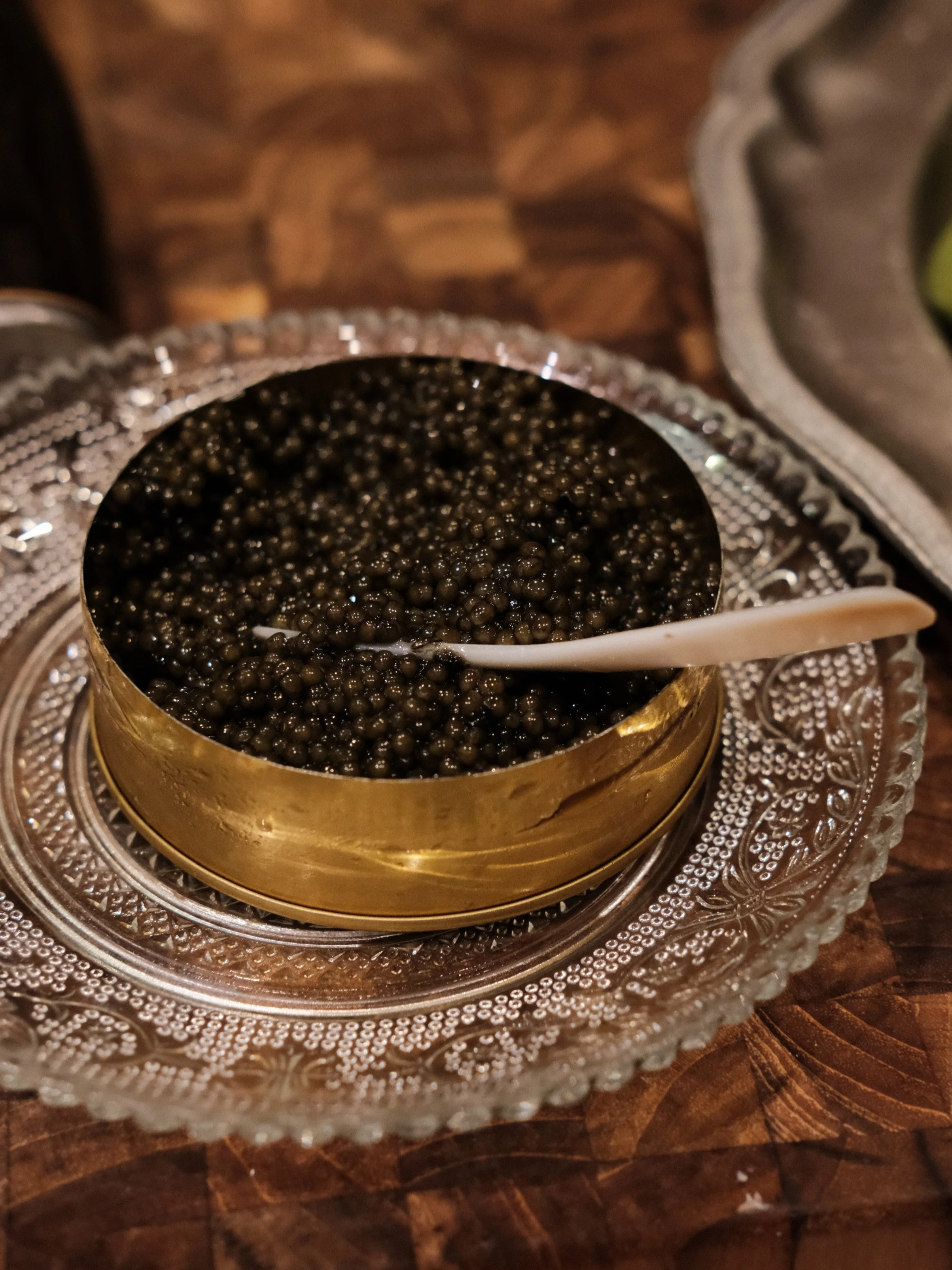 A bowl of black caviar with a small white serving spoon, placed on a decorative glass tray on a wooden surface.