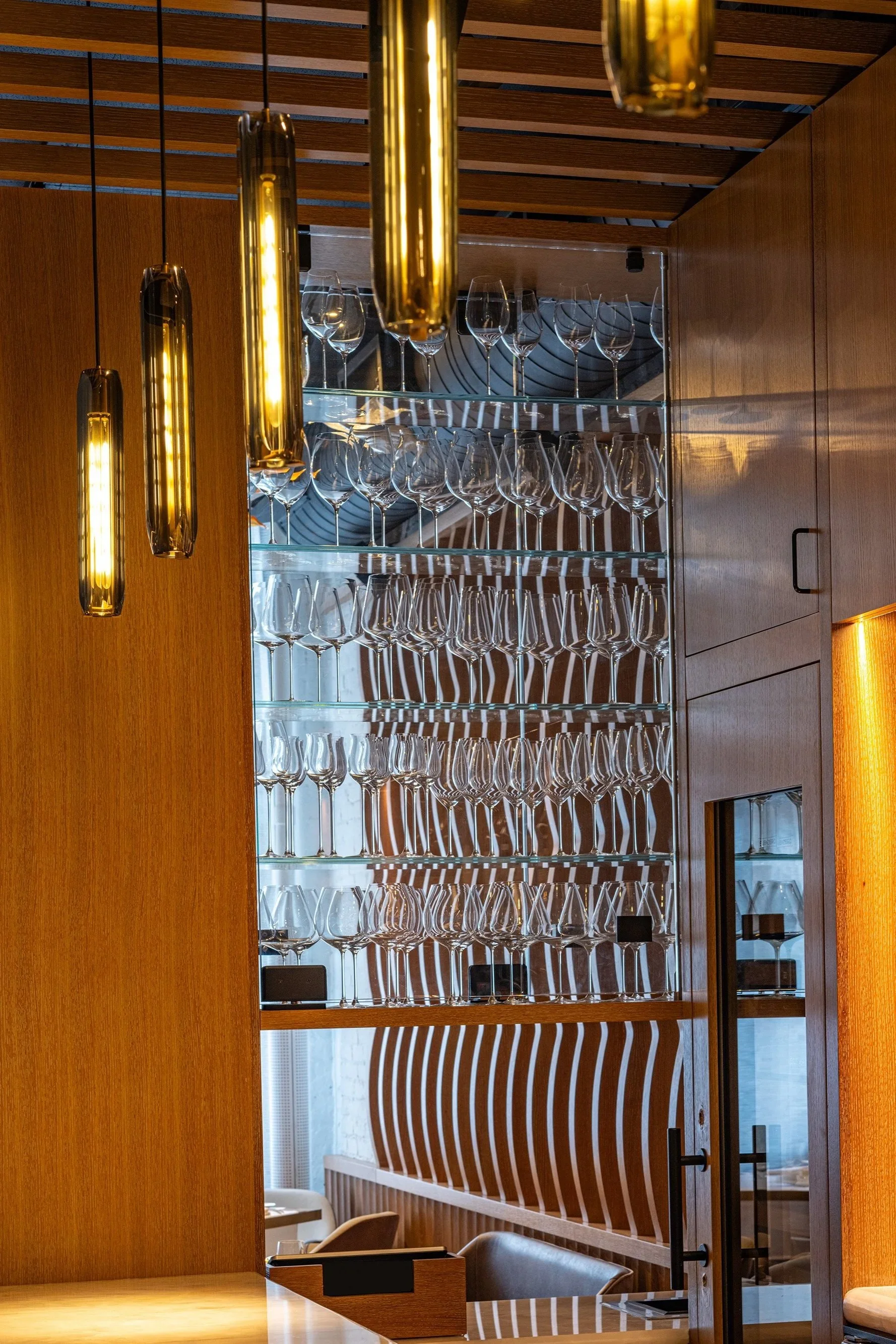 A display of empty wine glasses arranged on glass shelves in a modern restaurant, with warm wooden paneling and hanging gold-colored pendant lights.
