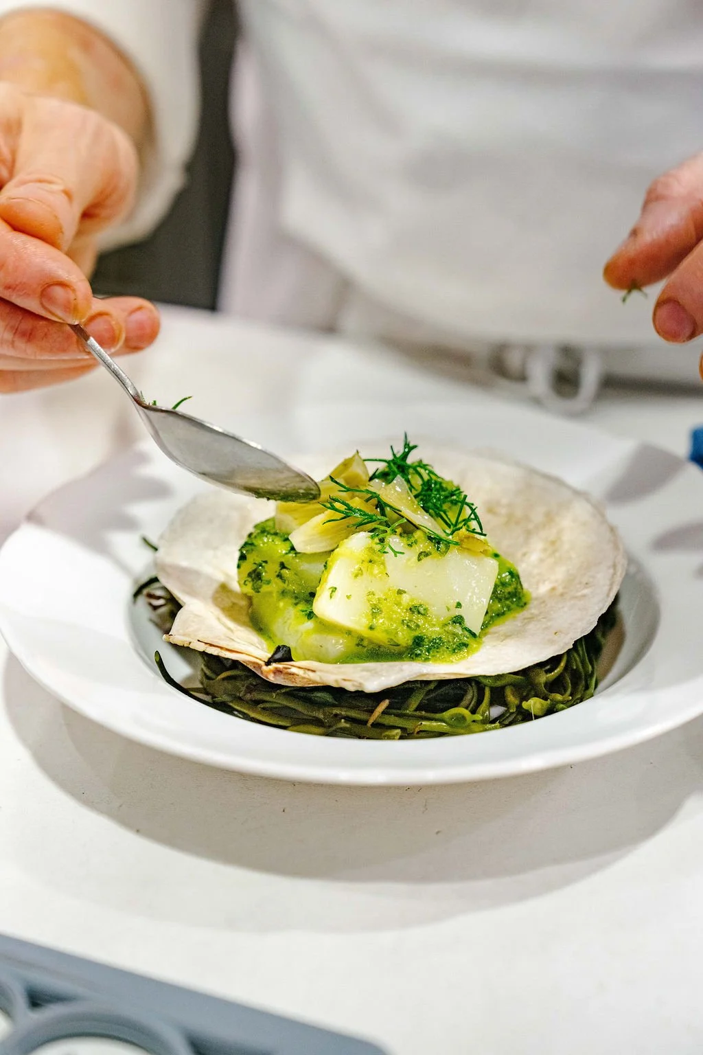 Person adding green herbs to a seafood dish with broccoli and potato in a shell on a white plate.