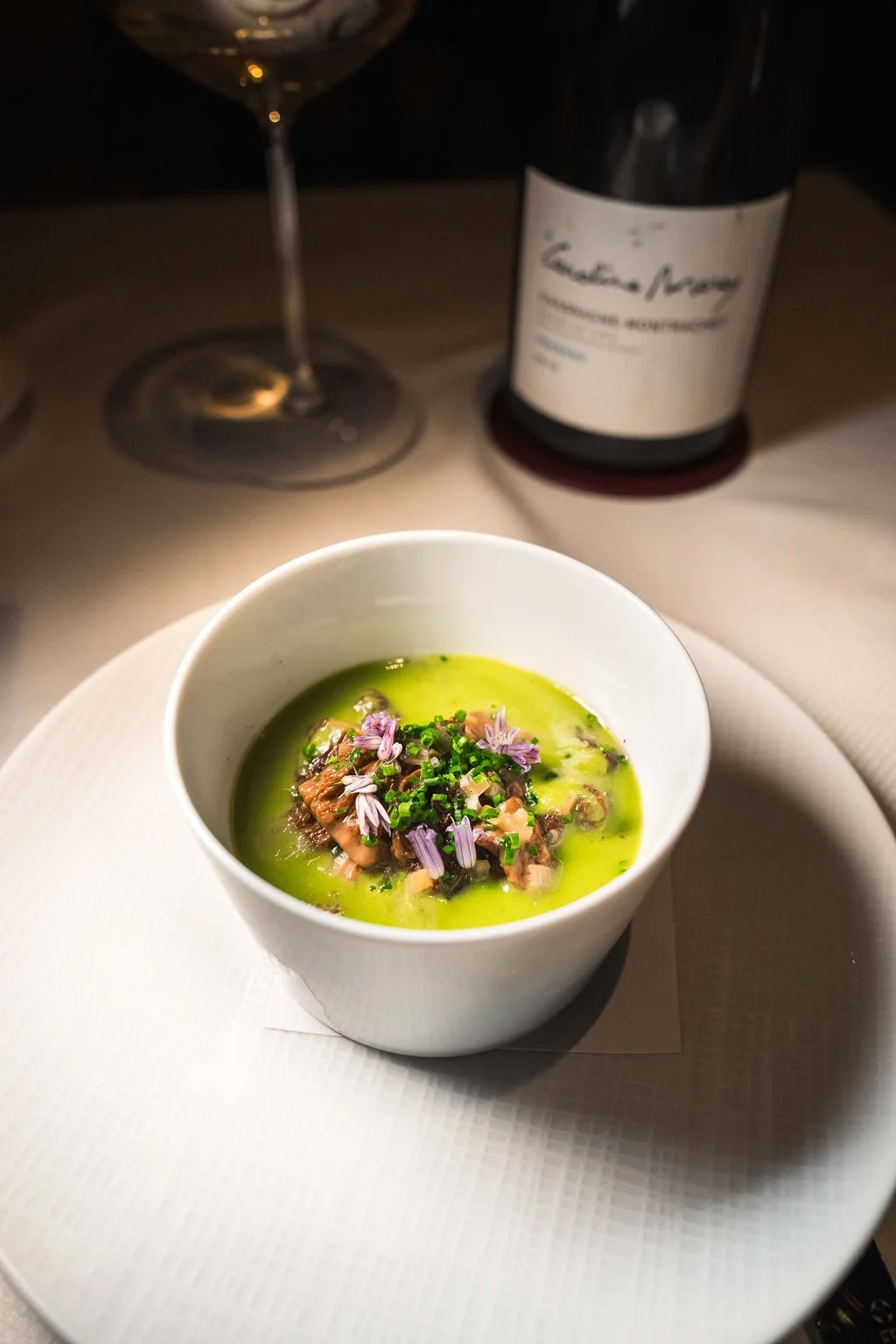 A white bowl of green soup garnished with chopped herbs and small purple edible flowers, served on a white plate. A glass of white wine and a bottle of wine are visible in the background.
