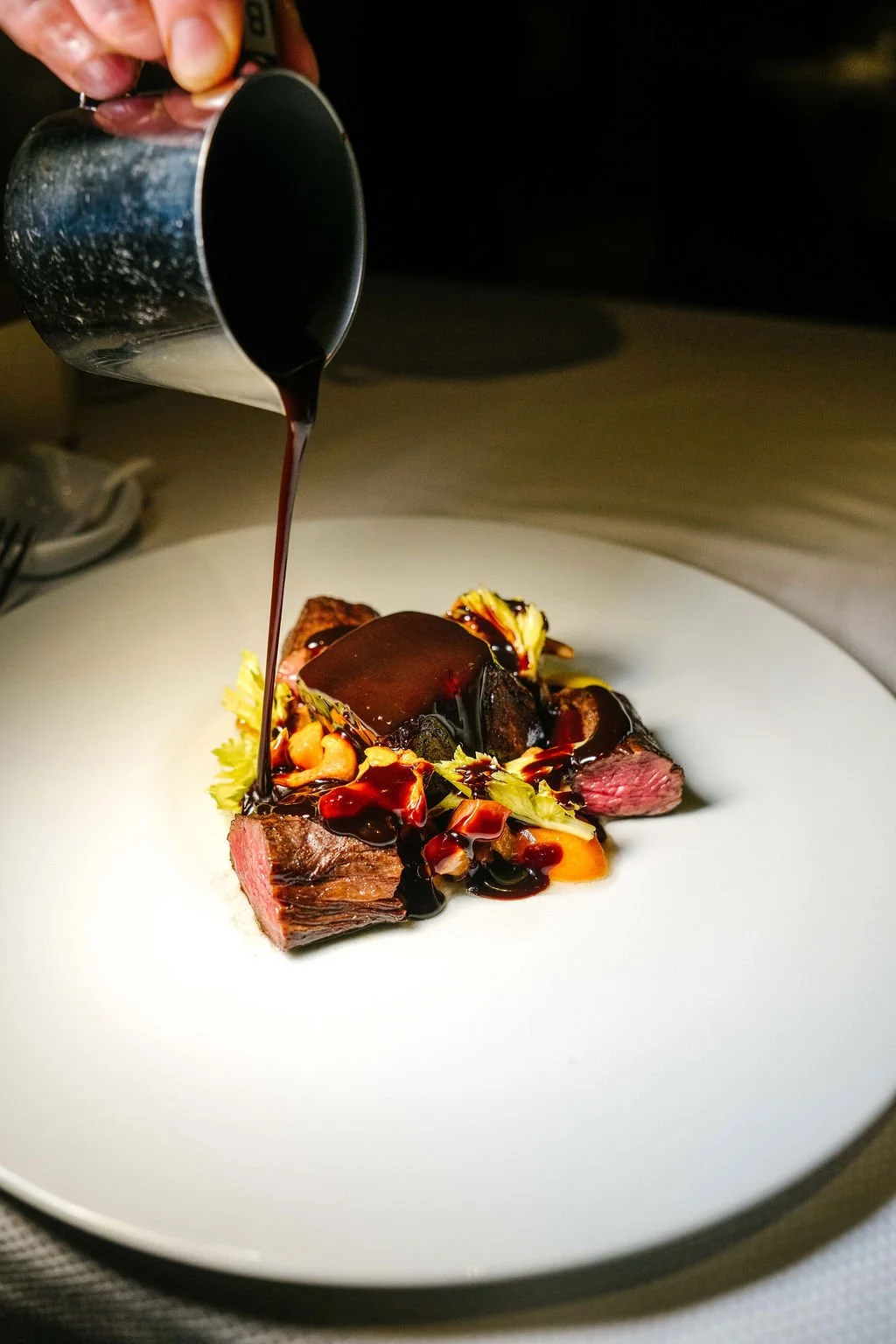A plate of medium-rare steak topped with dark sauce being poured over it, garnished with vegetables and a rich sauce.