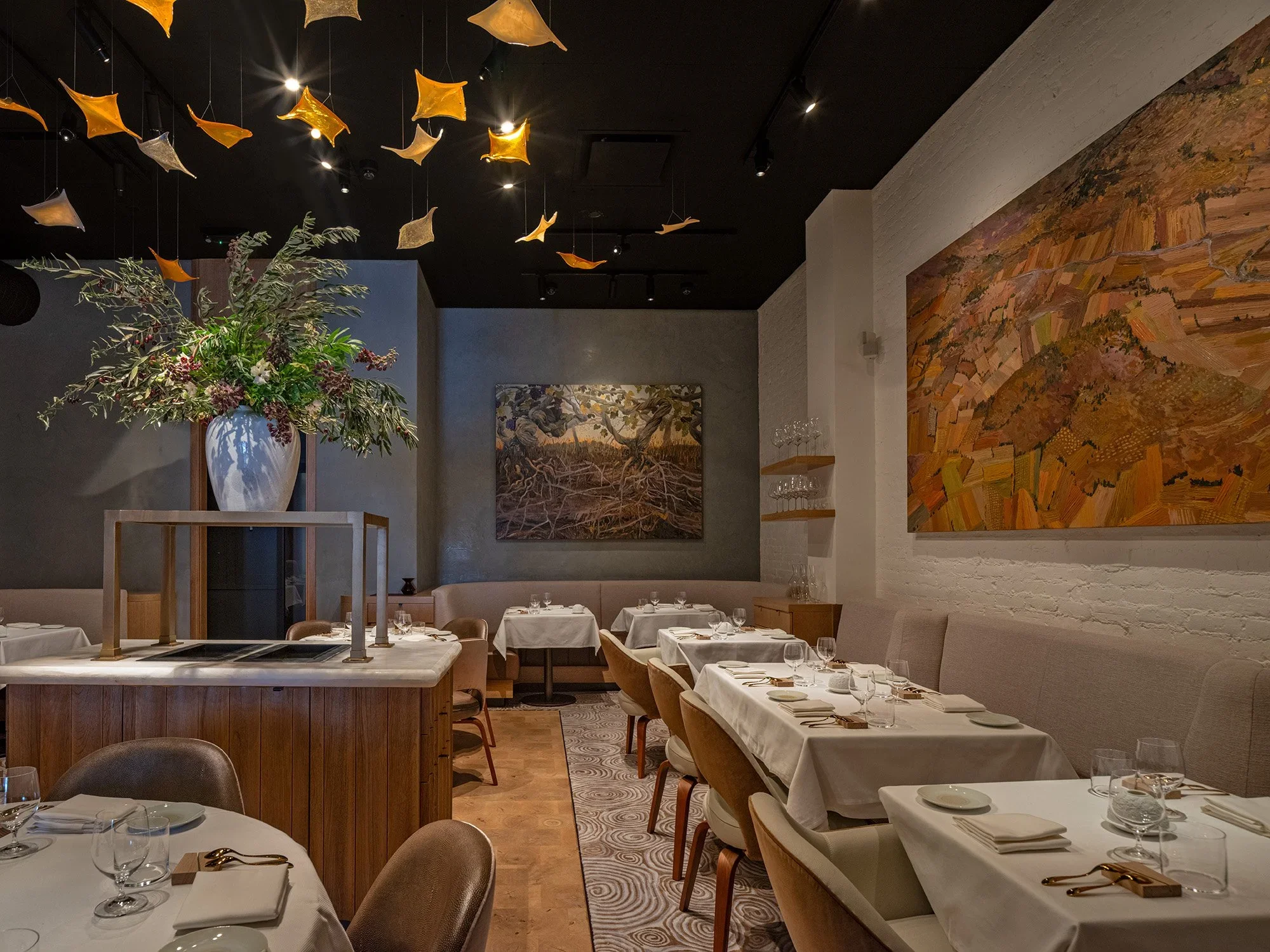 Elegant restaurant interior with white tablecloths, glassware, and gold utensils. Large floral arrangement with green foliage and purple berries in a white vase. Contemporary wall art and hanging yellow and beige fabric decorations. Soft lighting and