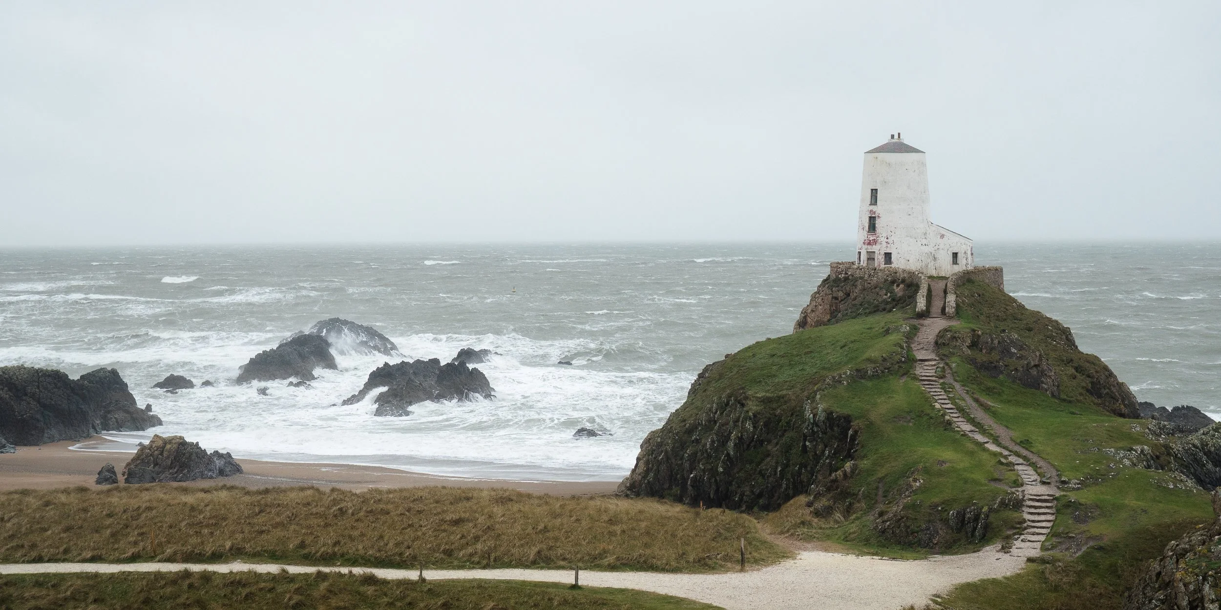 A lighthouse situated on a grassy hill overlooking the ocean, with rocky outcroppings in the water and a sandy beach in the foreground, under a cloudy, overcast sky.