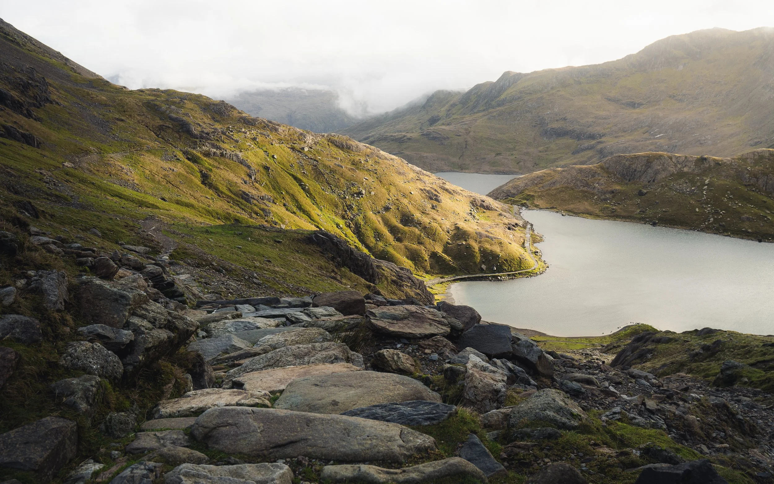 Scenic view of a mountain landscape with a rocky foreground, lush green slopes, and a winding lake surrounded by misty mountains.