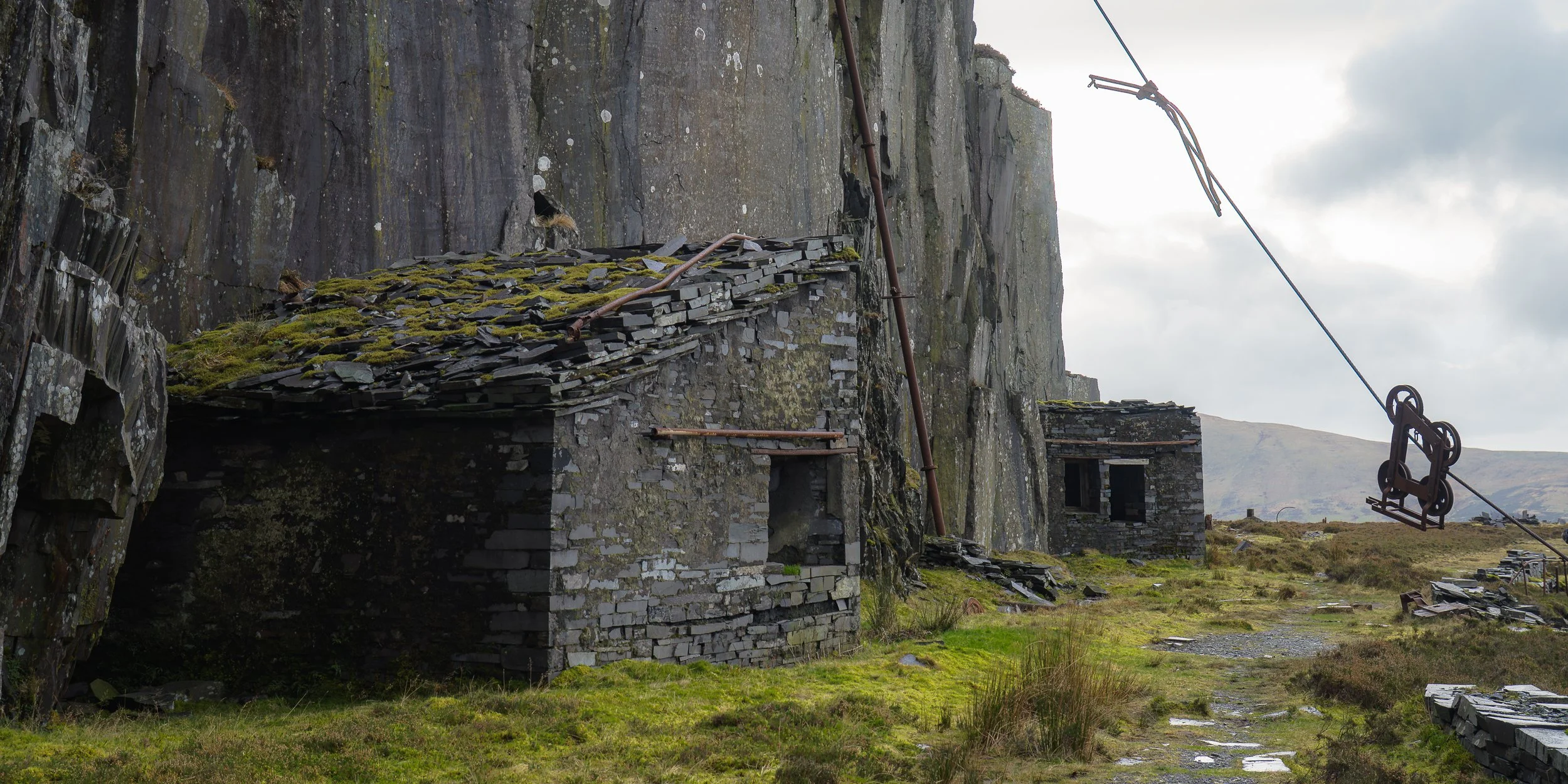 Abandoned stone buildings and rusted ski lift seats on a mountainous landscape under cloudy sky.