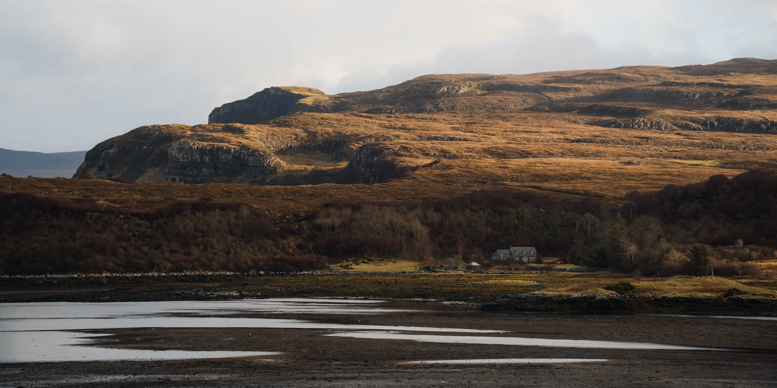 A scenic landscape featuring a body of water in the foreground, rolling brown hills, and rocky mountains in the background, with a house nestled among trees on the right side.