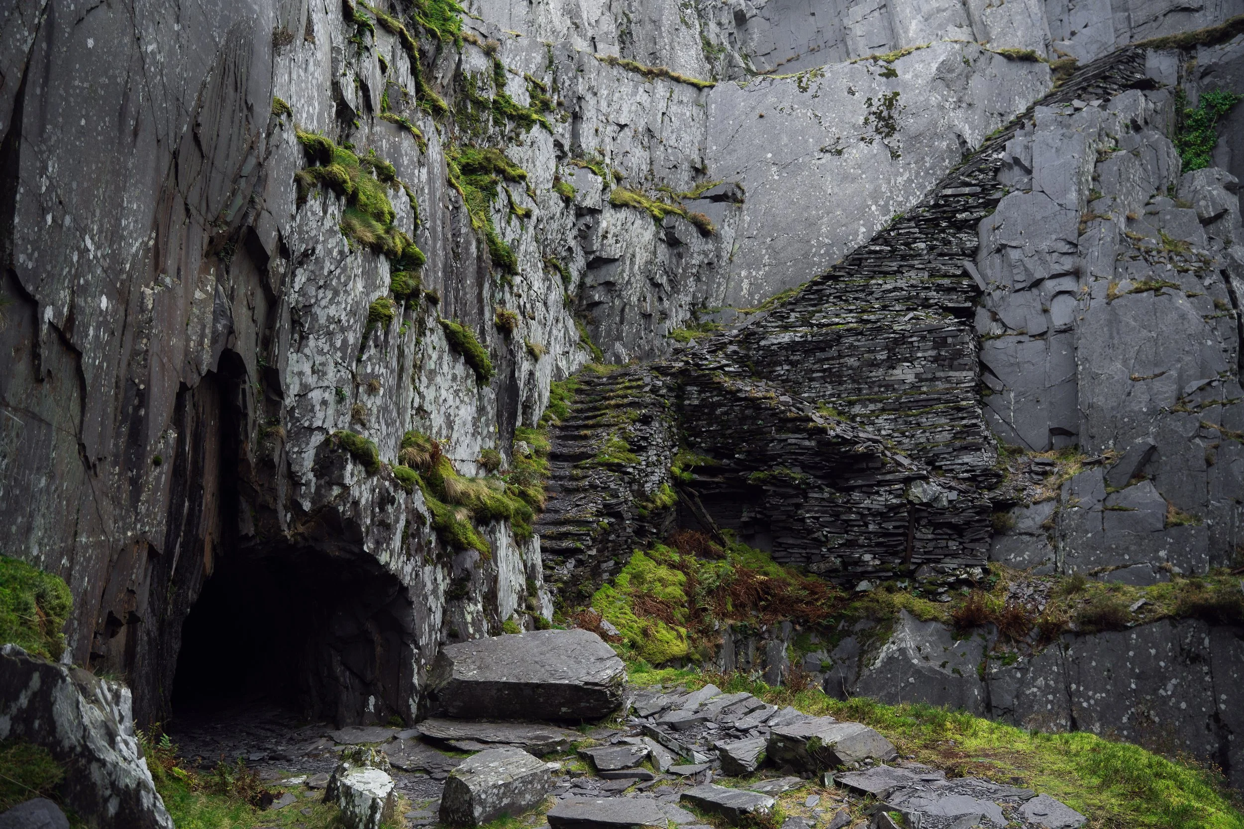 A rocky mountain landscape with moss-covered rocks, cliffs, and a small cave at the bottom left of the image.