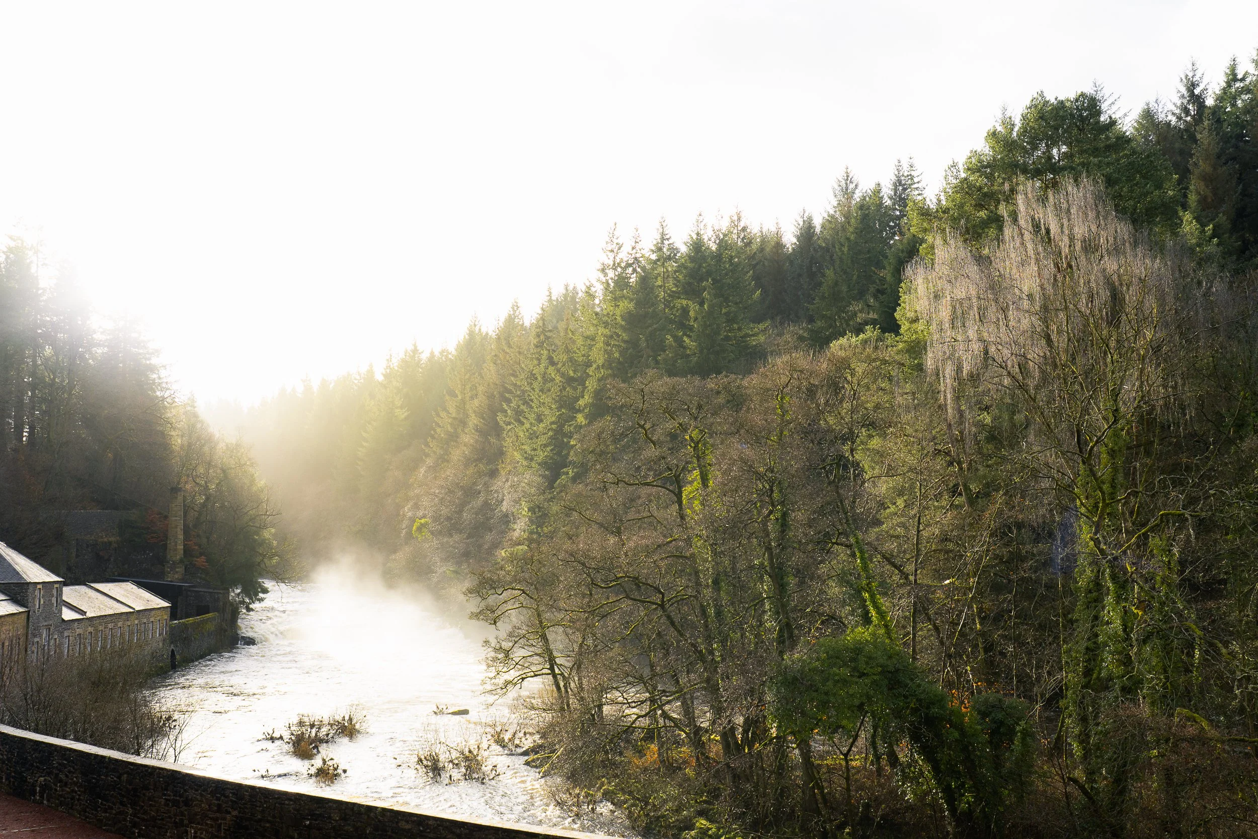 Sunlight shining over a misty river flowing through a forested landscape with trees and old buildings along the riverbank. Scotland.