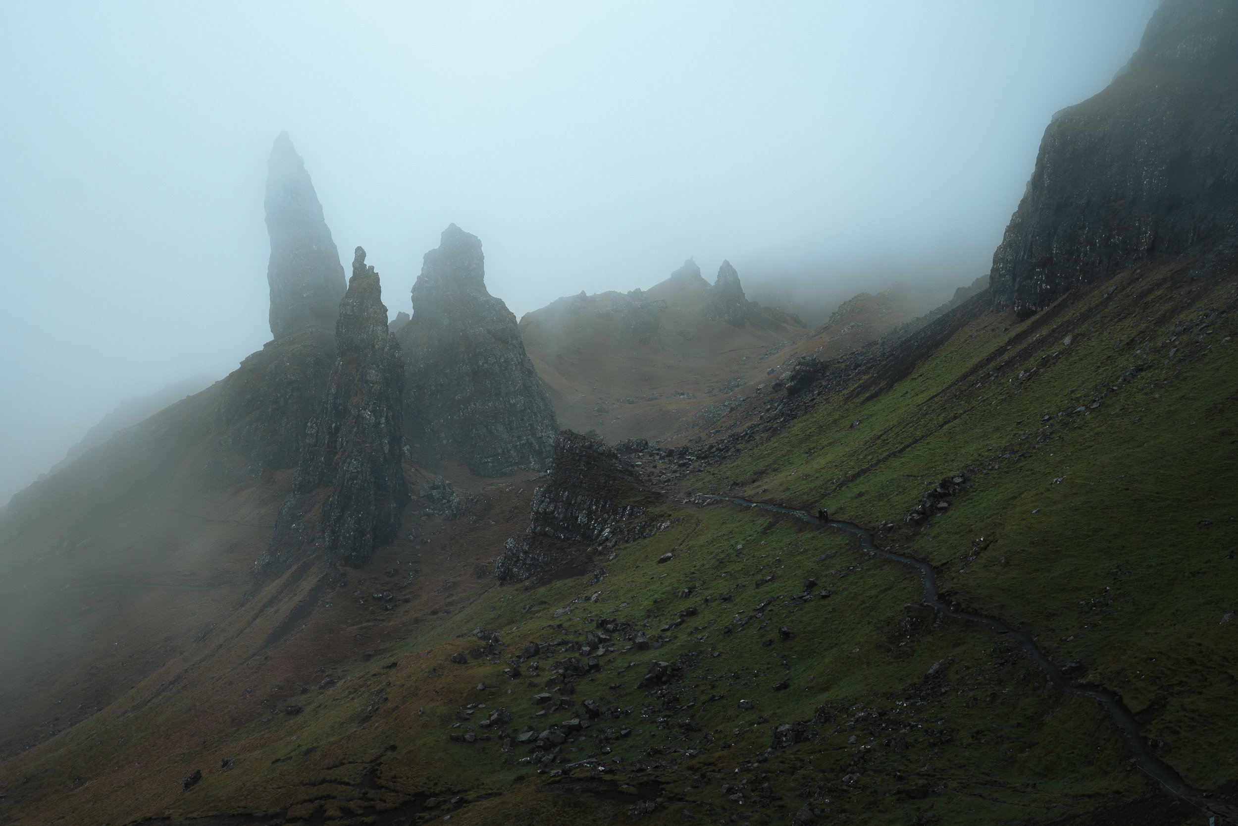 Mist-shrouded mountains with tall, jagged rock formations and a winding trail on a grassy hillside.