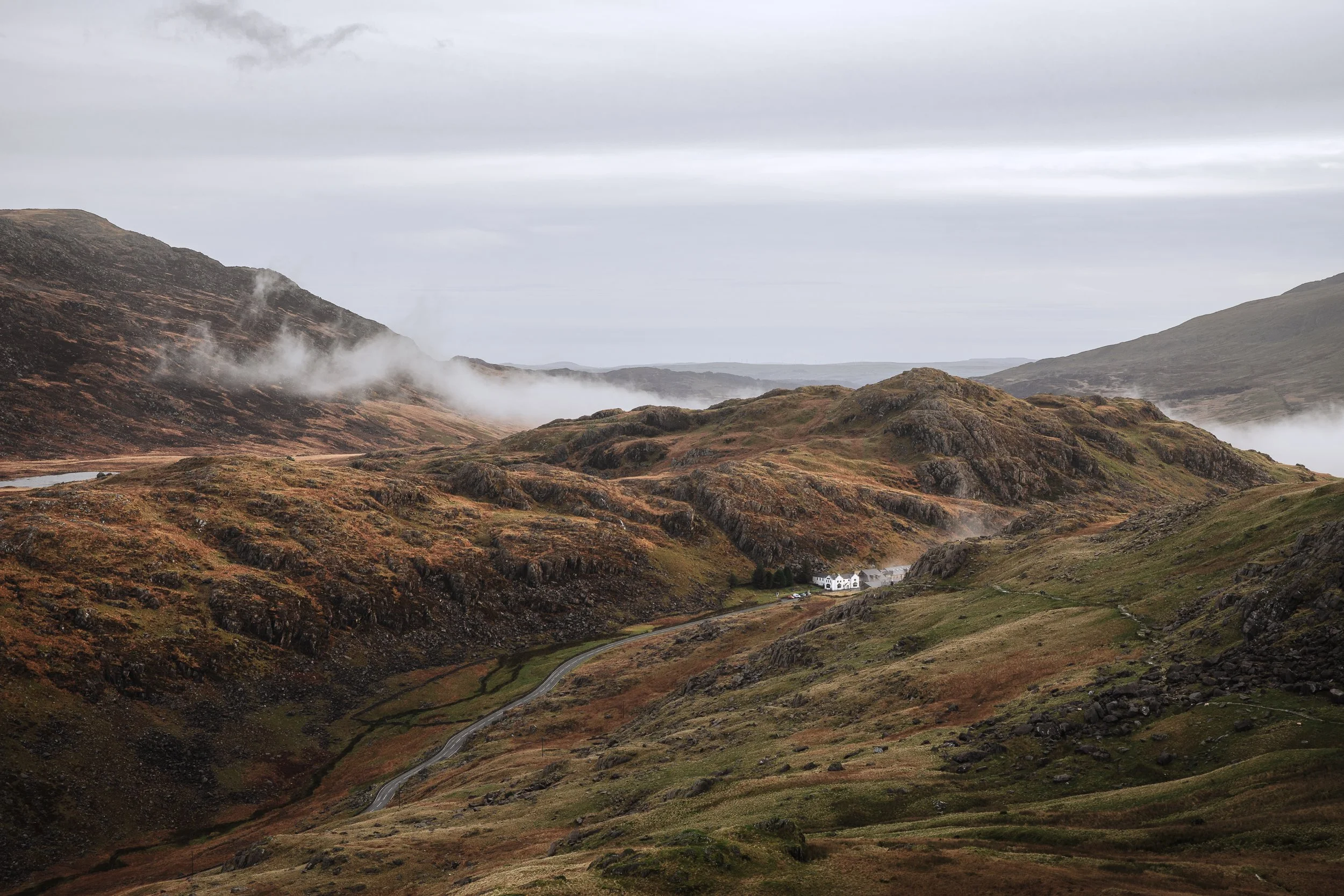 A scenic view of a cloudy, mountainous landscape with fog and a winding road leading to a small cluster of white buildings.