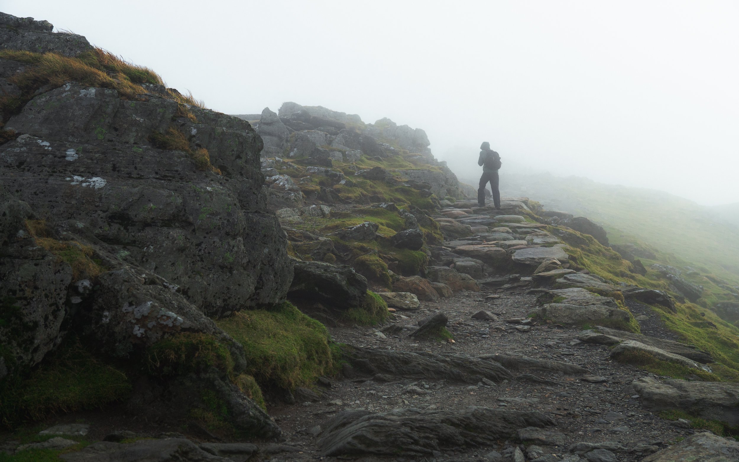 A lone hiker with a backpack walking along a rocky mountain trail shrouded in fog. Snowdon, North Wales.