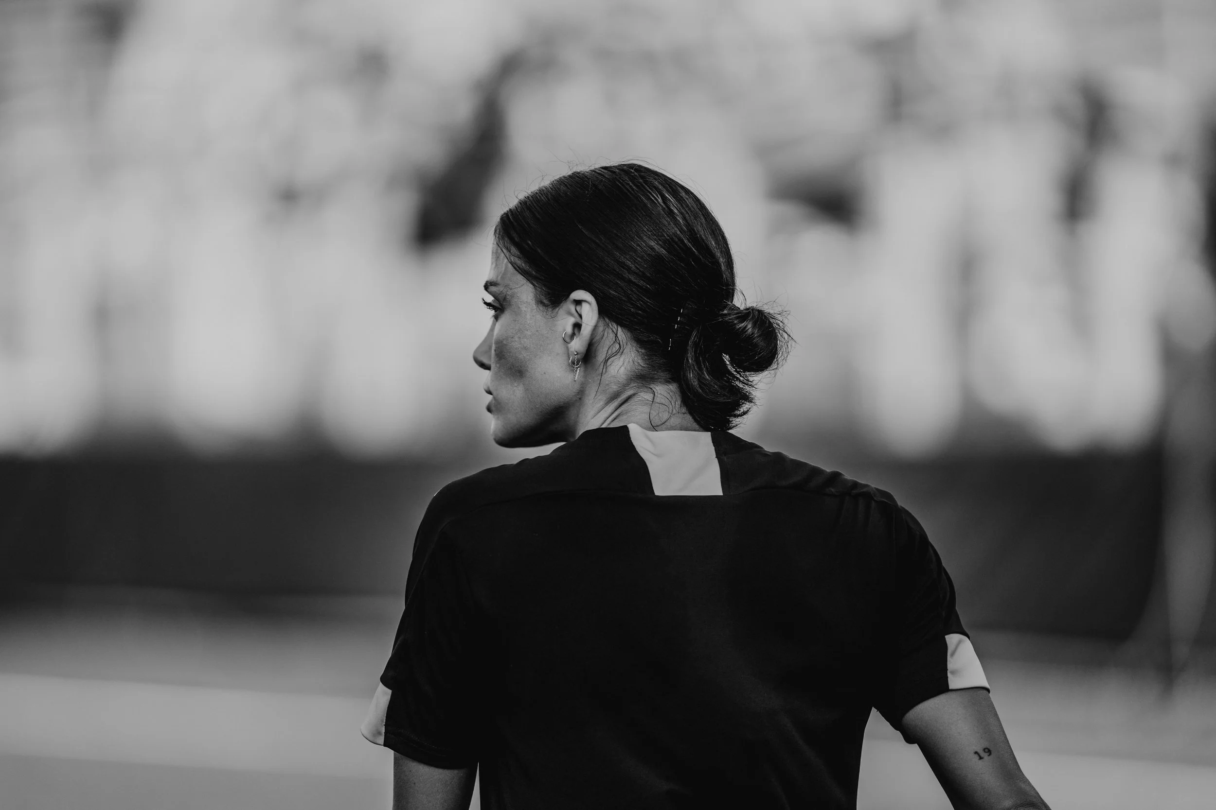 Black and white photo of a woman with dark hair in a low bun, wearing a sports jersey, standing outdoors with a blurred background.