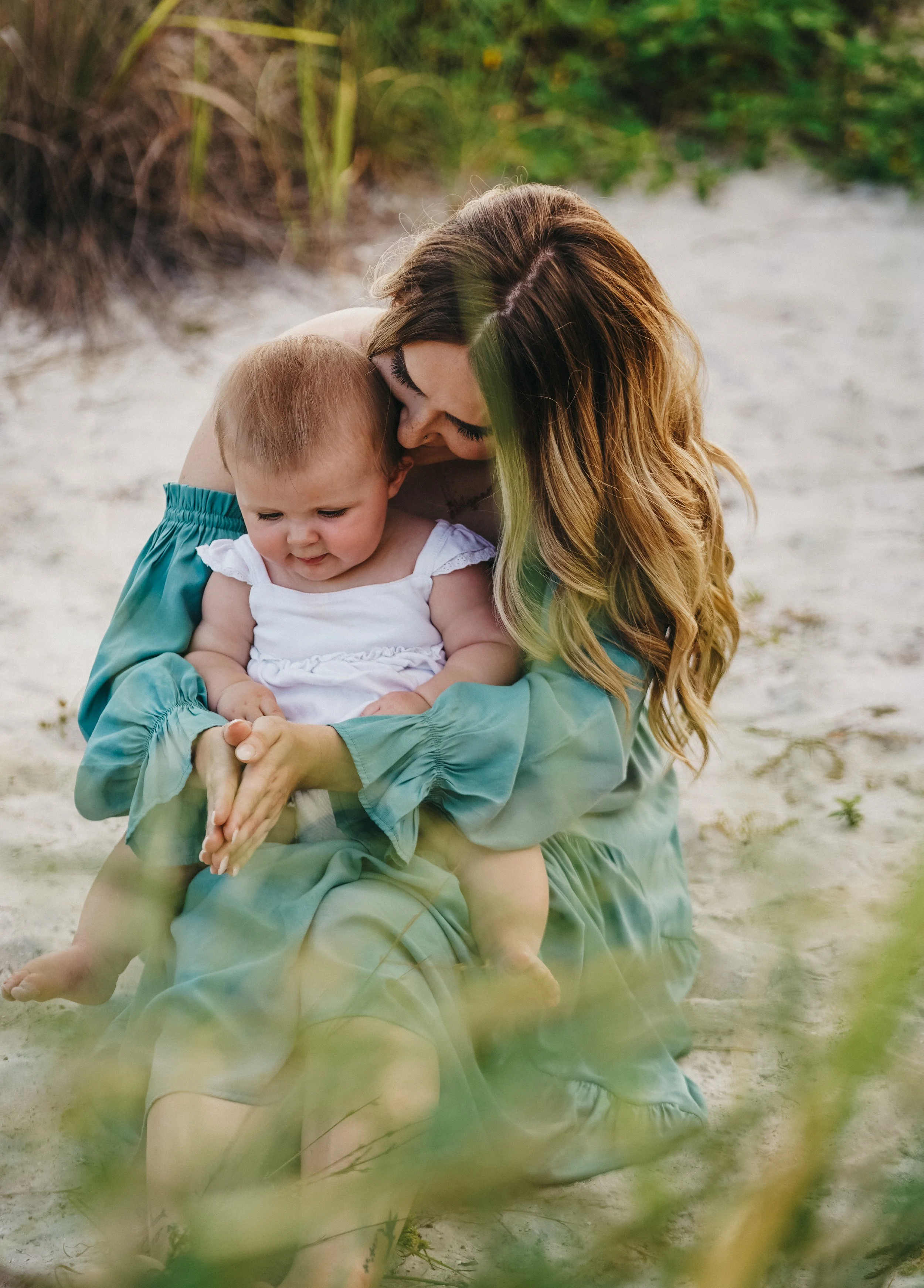 A woman with long wavy hair holding a young girl on a sandy beach, with greenery in the background.