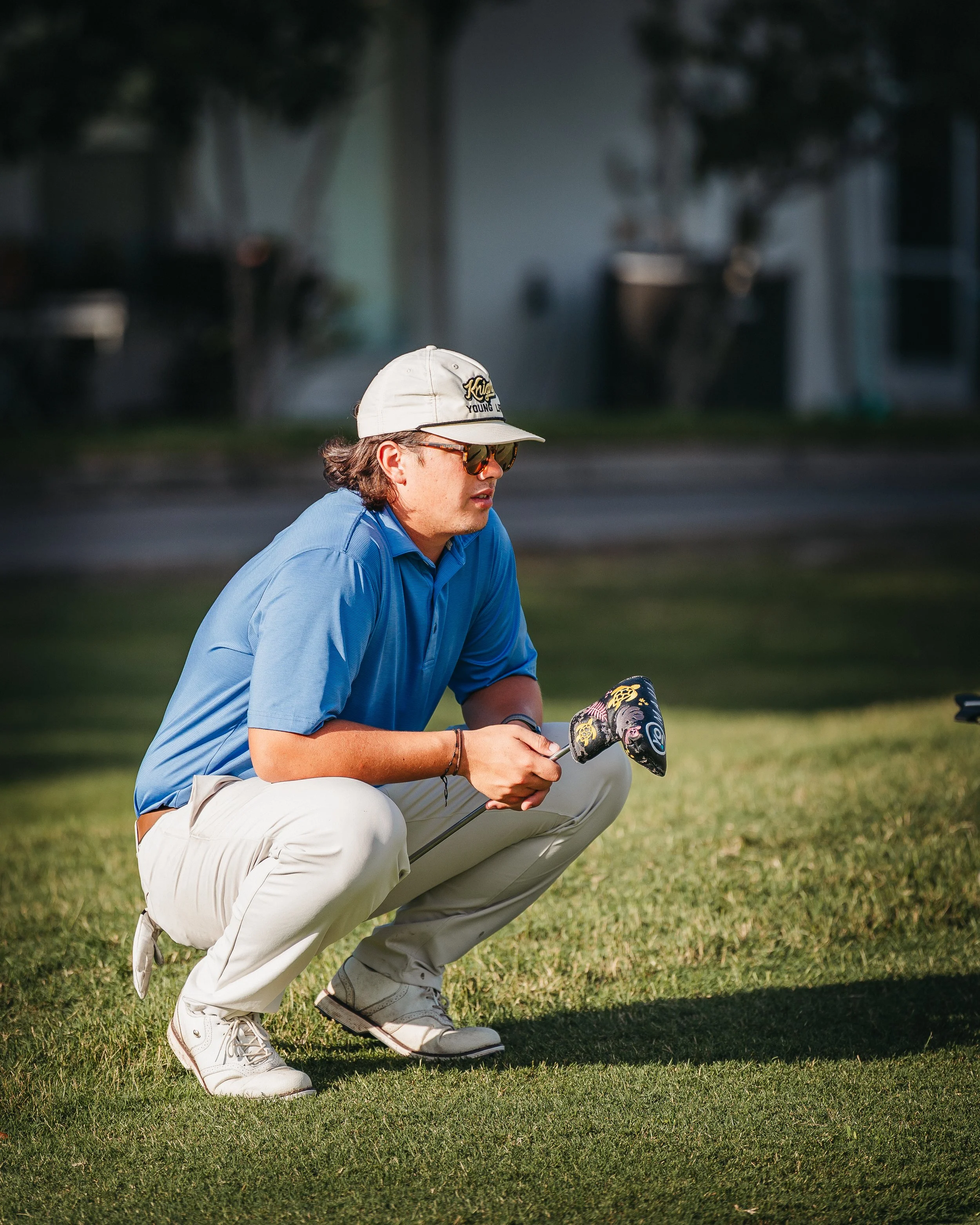 A man squatting on a golf course grass, holding a golf club, wearing a blue polo shirt, beige pants, white golf shoes, a gray cap, and sunglasses.