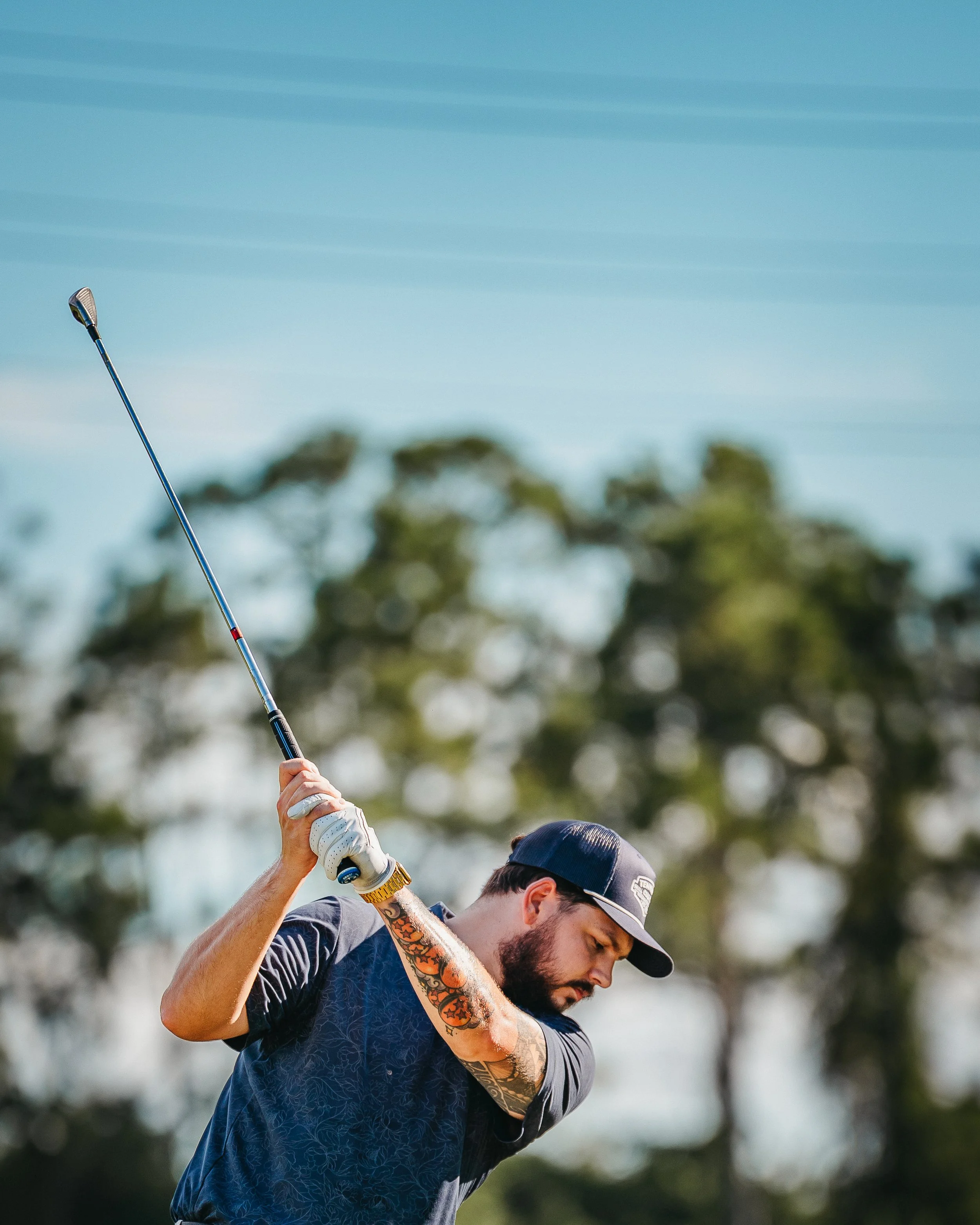 A man with tattoos on his arm wearing a dark cap and shirt swings a golf club on a golf course with trees in the background.