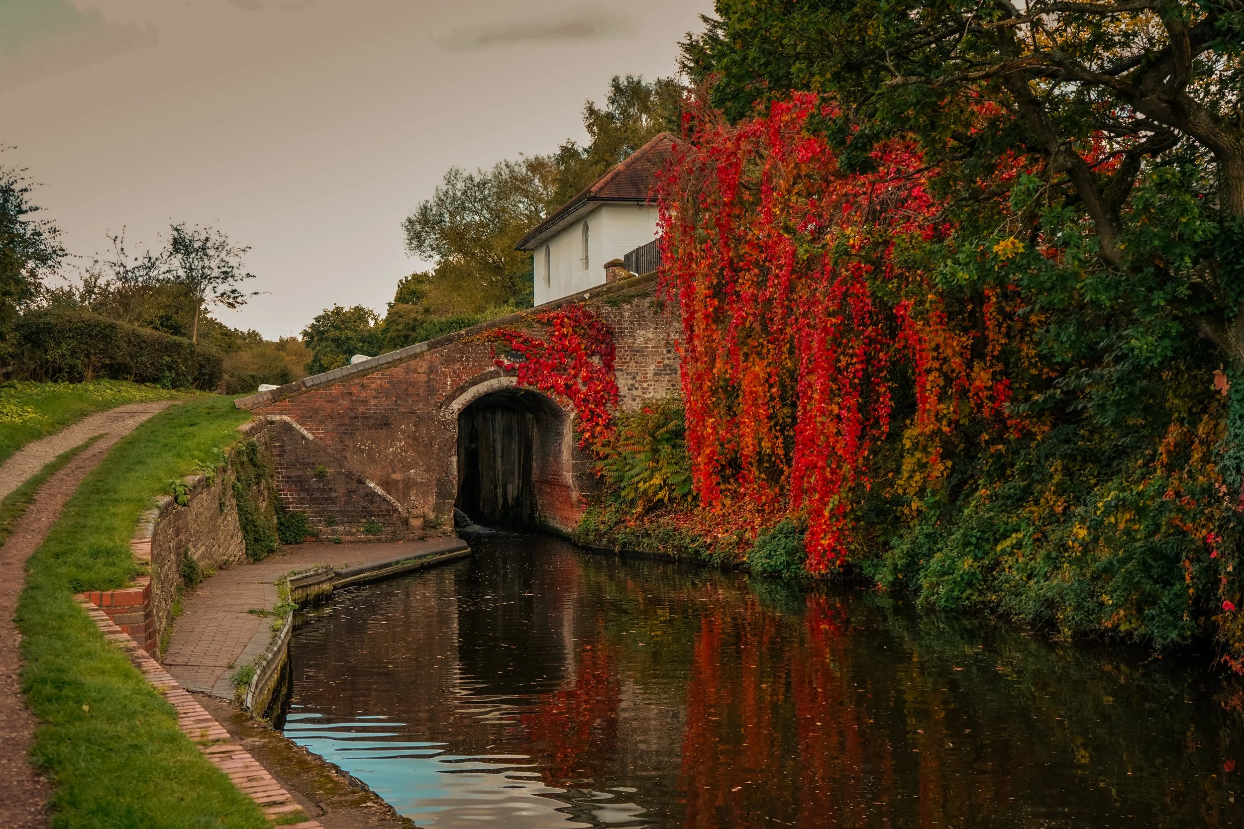 A historic brick and stone bridge over a narrow waterway with a white building on top, surrounded by trees with vibrant red, orange, and green autumn foliage.