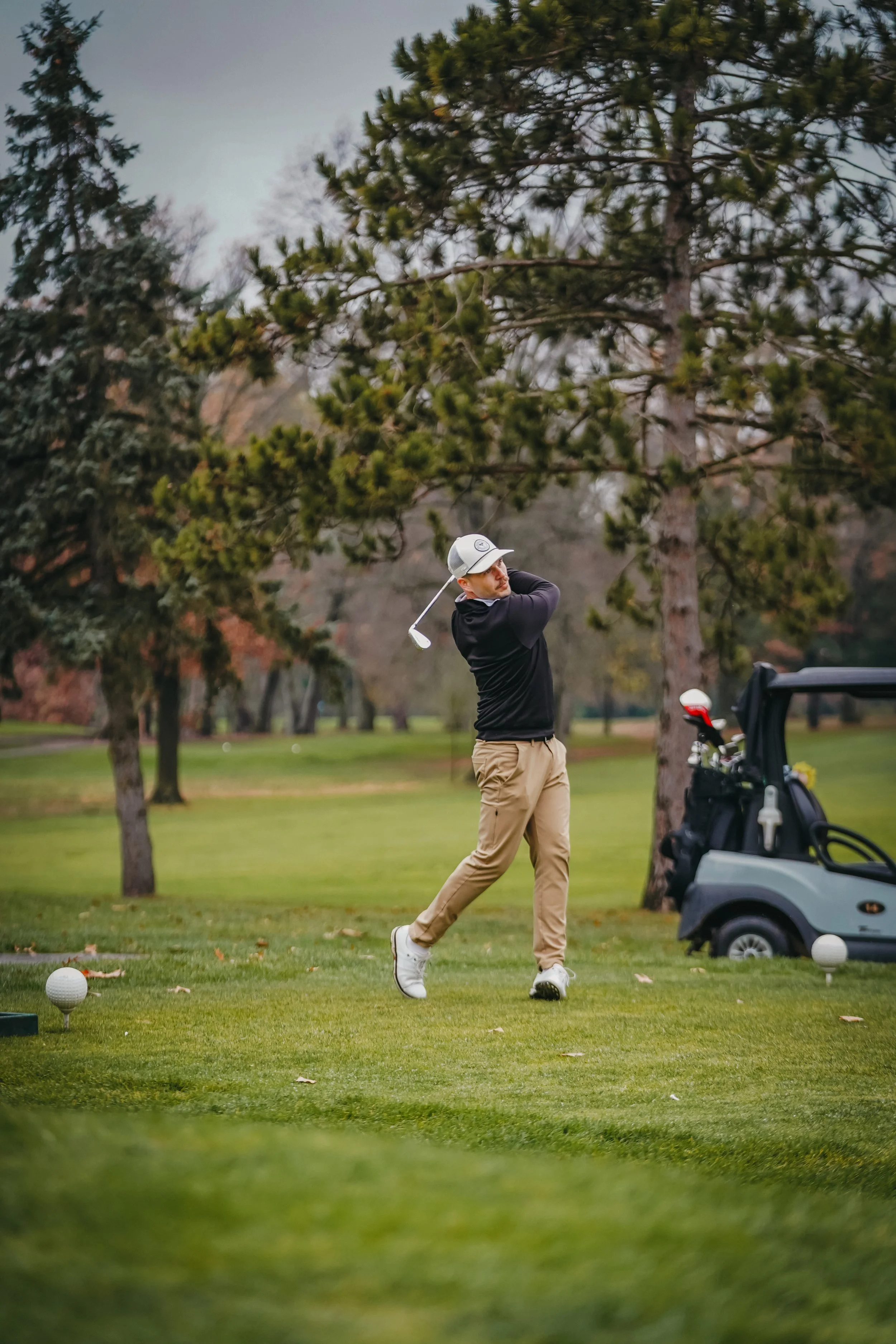 A man playing golf on a lush green course, swinging a club near a golf cart with trees in the background.