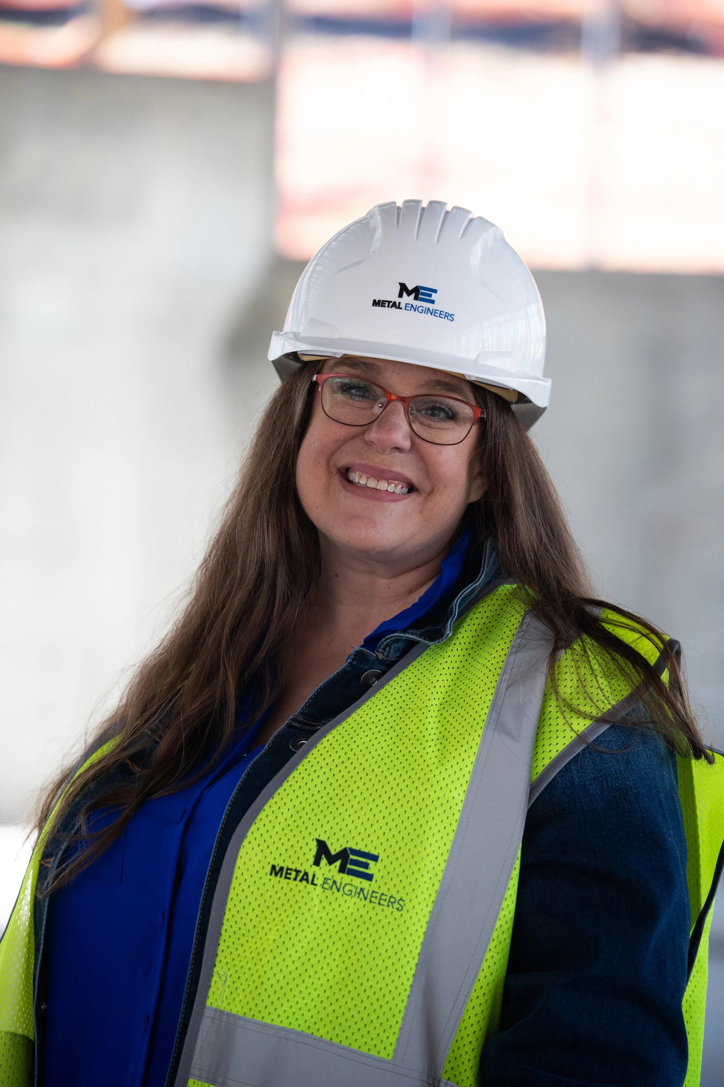 Woman in a blue shirt and yellow safety vest wearing a white hard hat with the Metal Engineers logo, smiling in an industrial setting.