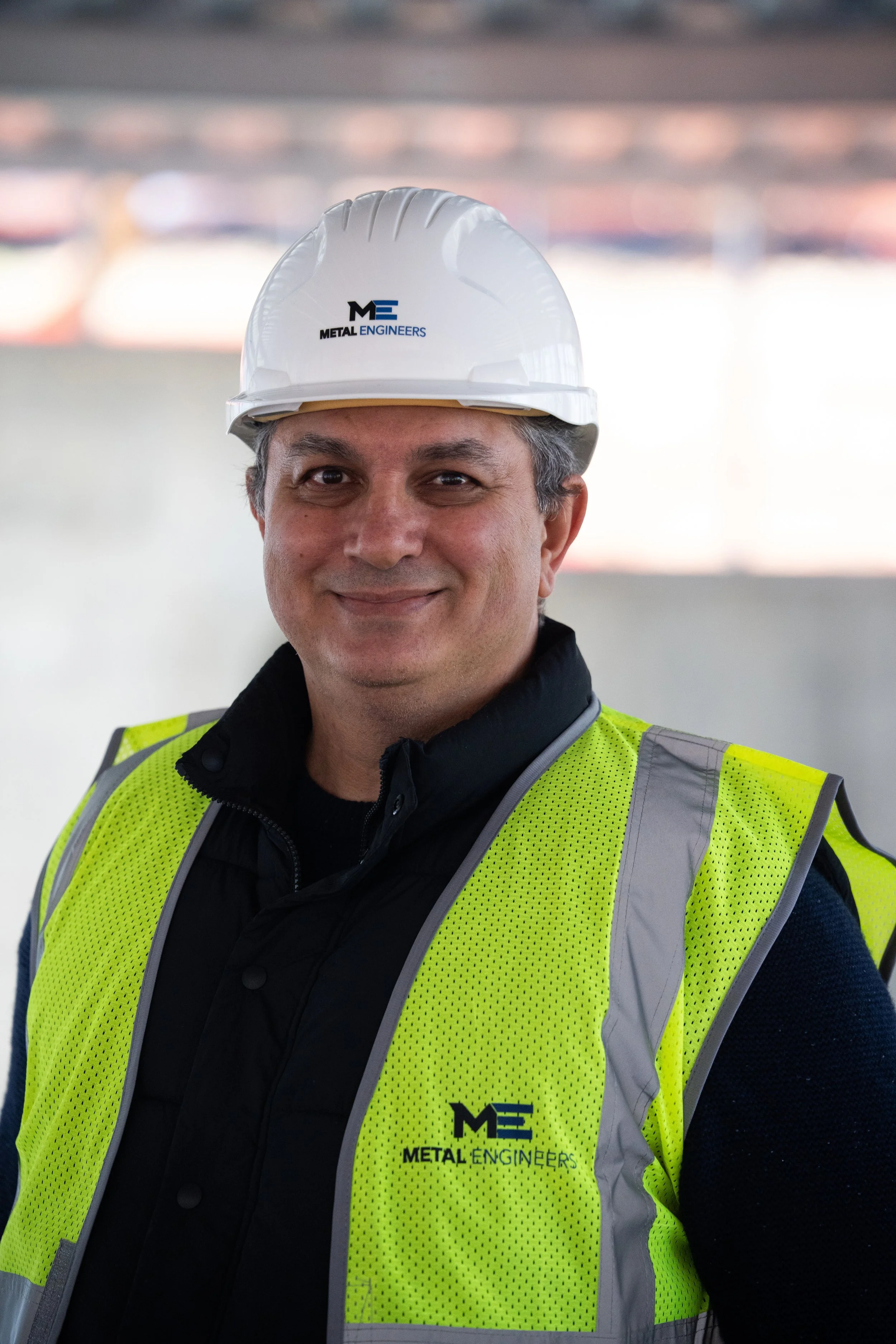 A man wearing a white hard hat and a yellow safety vest with the Metal Engineers logo, smiling at the camera.