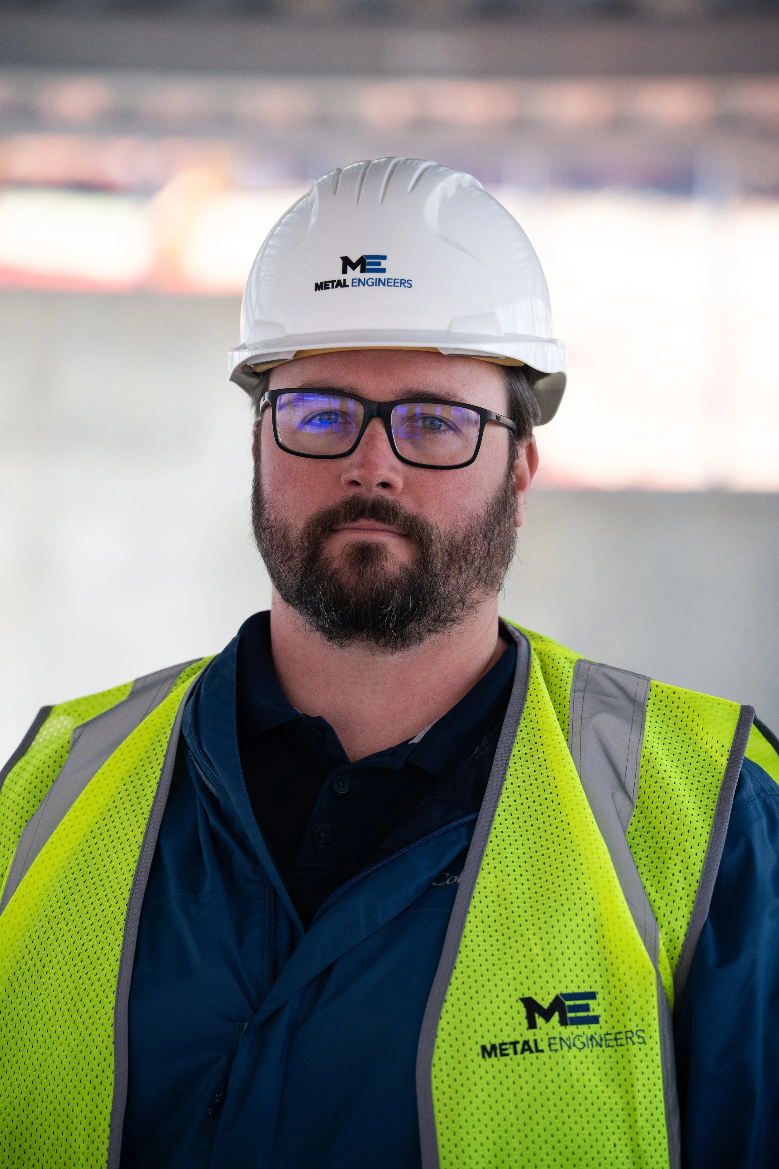 A man with a beard and glasses wearing a white safety helmet and a neon yellow safety vest with the logo 'Metal Engineers'.