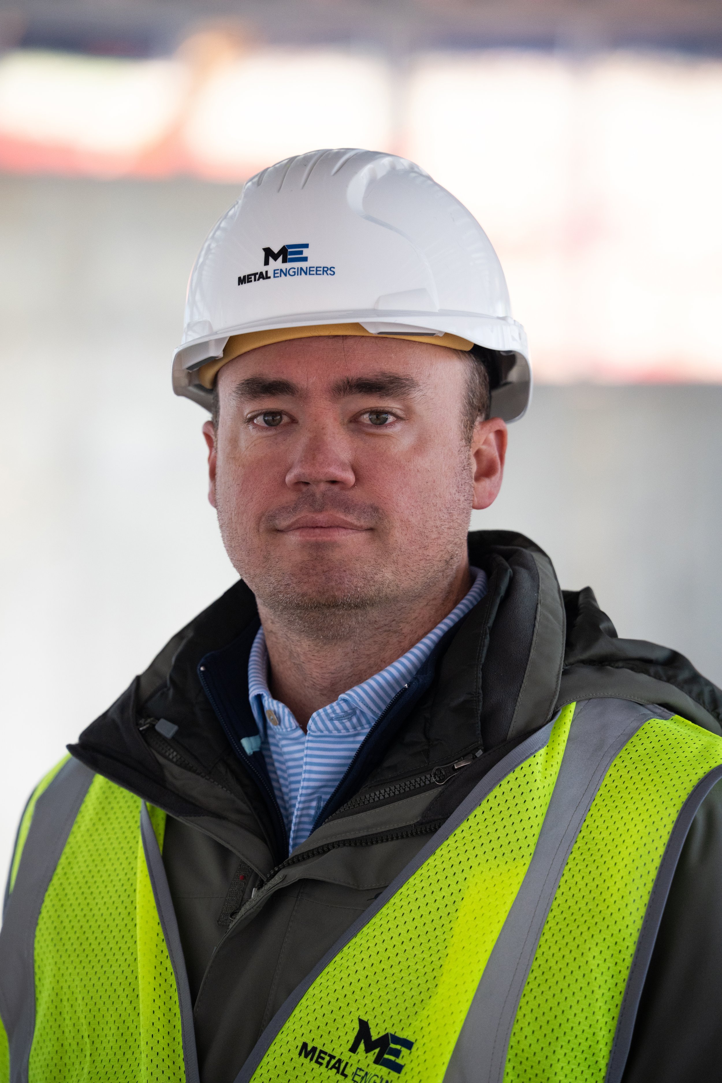 Man wearing a white hard hat and high-visibility safety vest with 'Metal Engineers' logo, indoors with blurred background.