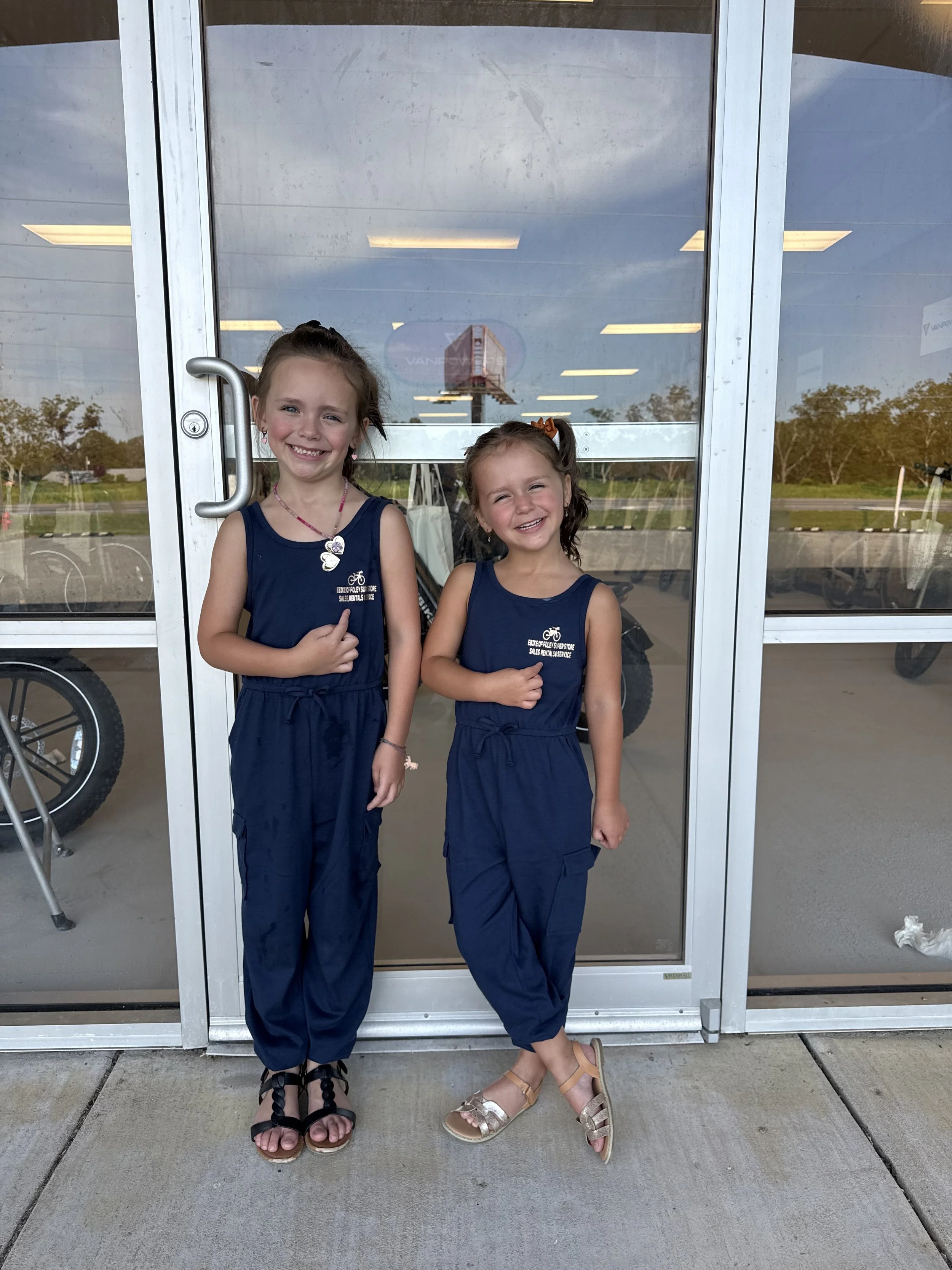 Two young girls in matching navy blue outfits standing outside in front of a glass door, smiling at the camera. They appear to be at a recreational or school event.