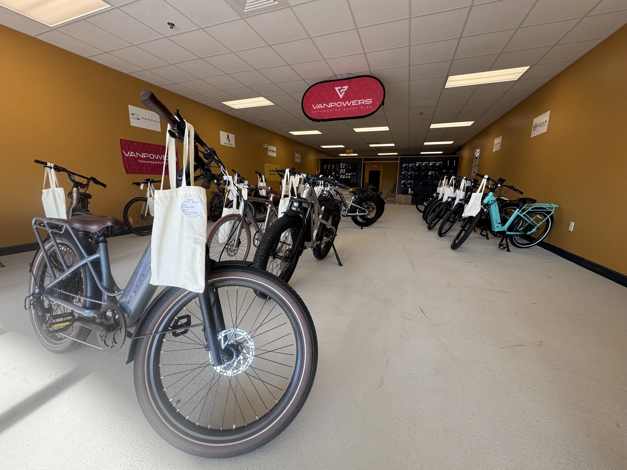 A room with multiple bicycles on display, some with tote bags hanging on the handlebars, and a pink Vanpowers sign hanging from the ceiling, with a yellow wall and ceiling panels.