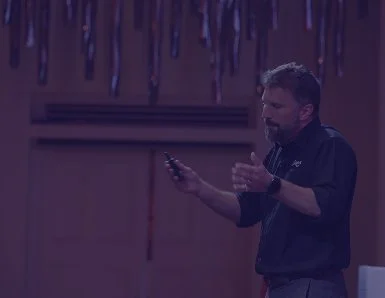 A man with gray hair and beard looking at his phone, standing in front of a wall decorated with hanging streamers.